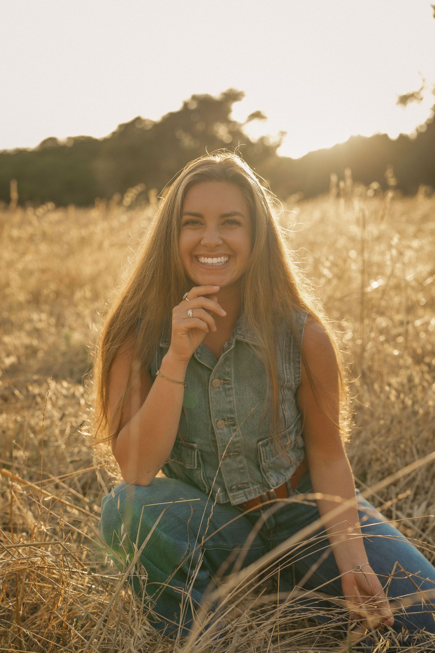 Smiling woman with long hair crouching in a golden grassy field at sunset, wearing a denim vest and jeans.