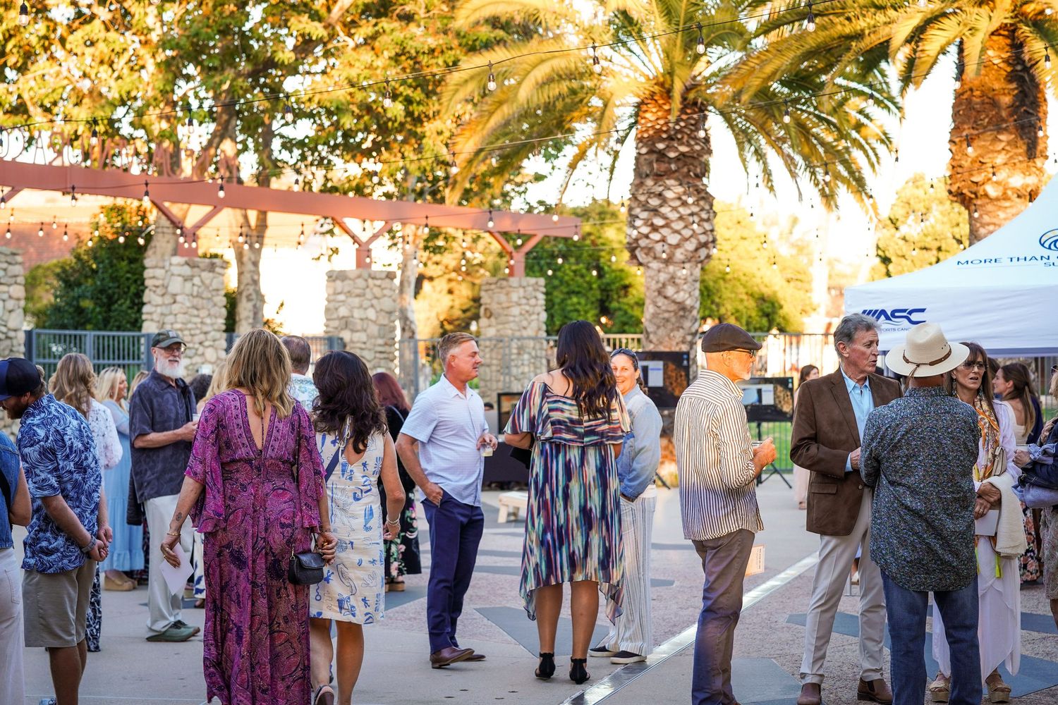 Guests mingle outdoors under palm trees near a stone archway and tent at an evening event.