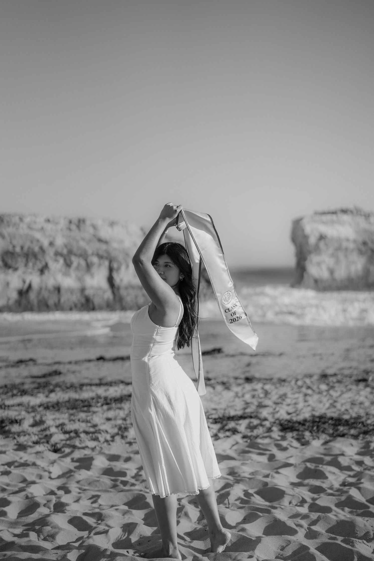 Black and white photo of a woman in a white dress holding a sash above her head on a beach with rock formations.