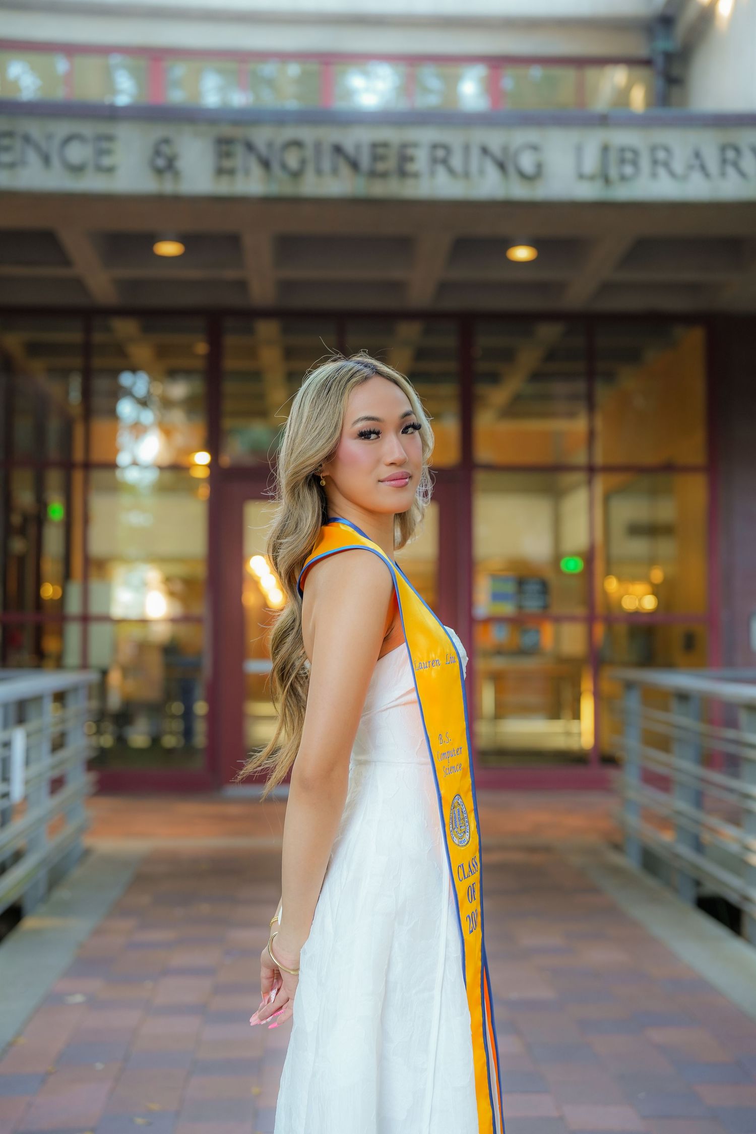 Graduate in a white dress and gold stole stands in profile on a brick walkway outside the Science and Engineering Library.