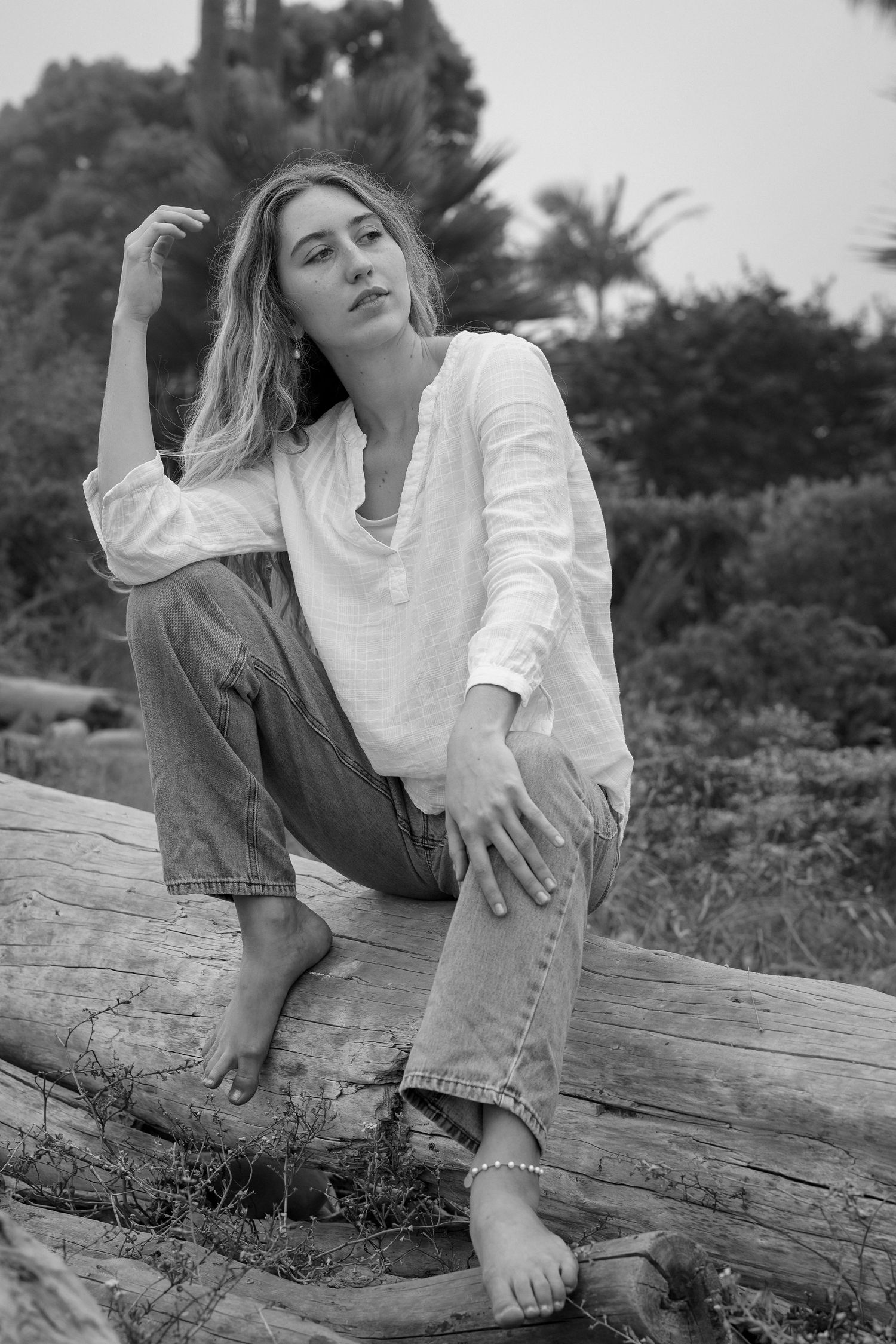 Black and white portrait of a woman in a loose white shirt and jeans sitting barefoot on a driftwood log, gazing into the distance.