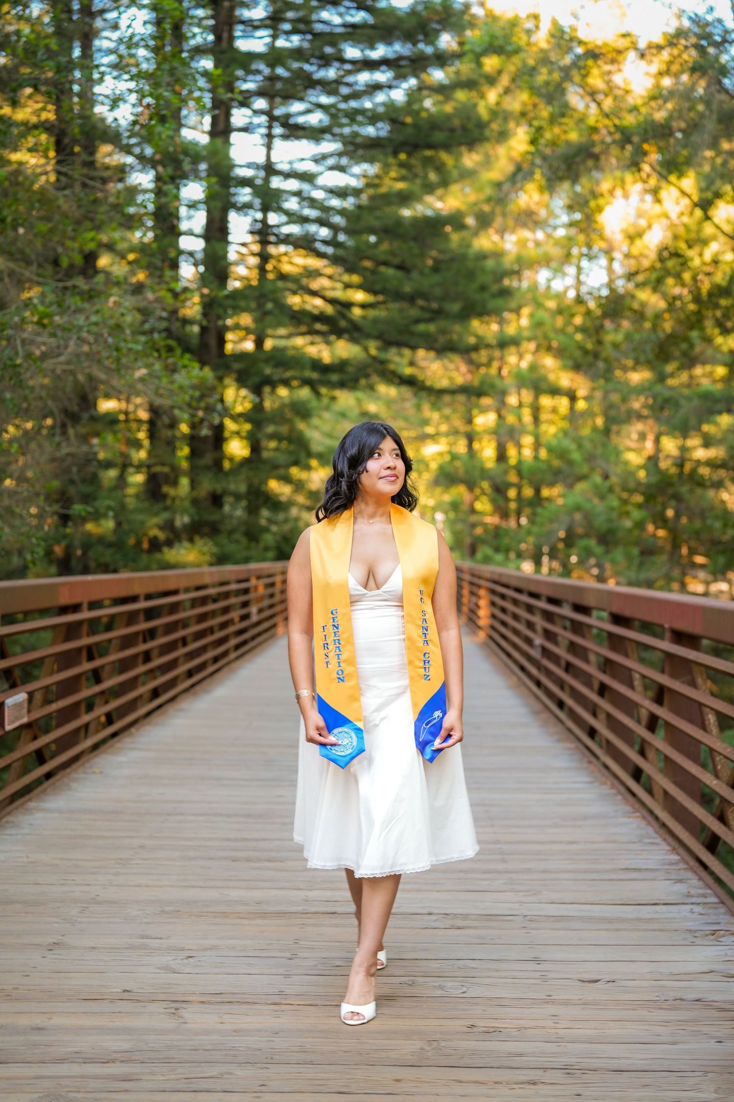 Graduate in a white dress and yellow stole walking across a wooden bridge surrounded by sunlit forest trees.