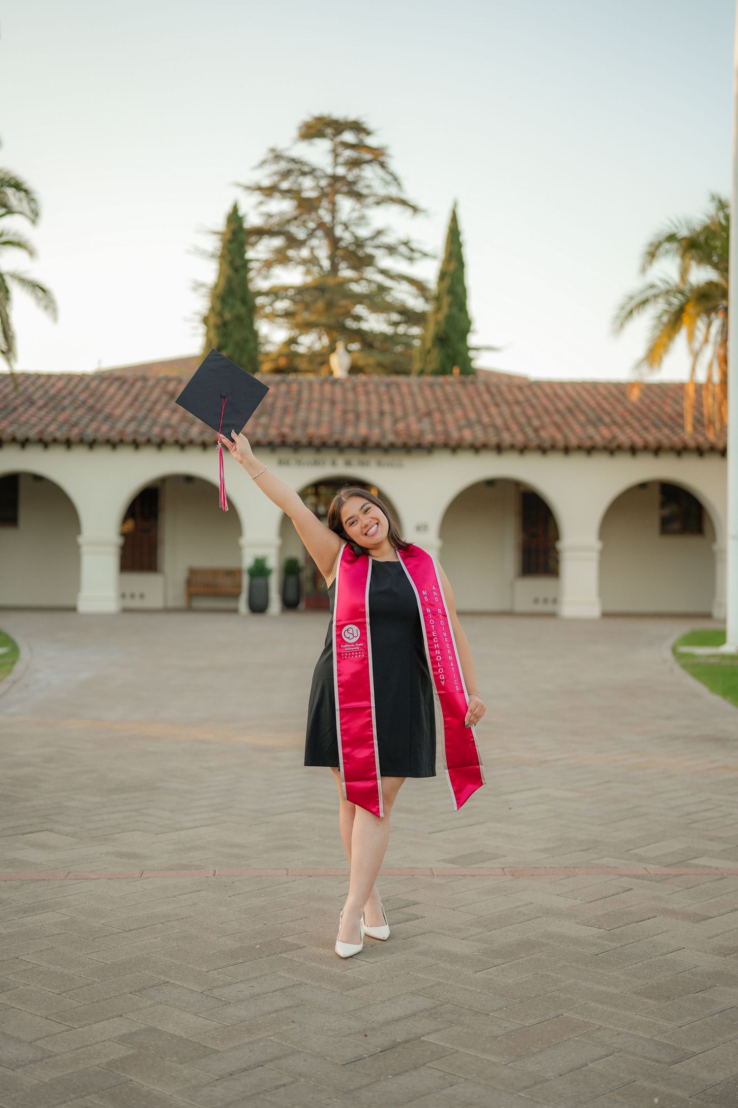 Graduate in a black dress and pink stole raises her cap overhead while standing on a brick courtyard before a Spanish-style building.