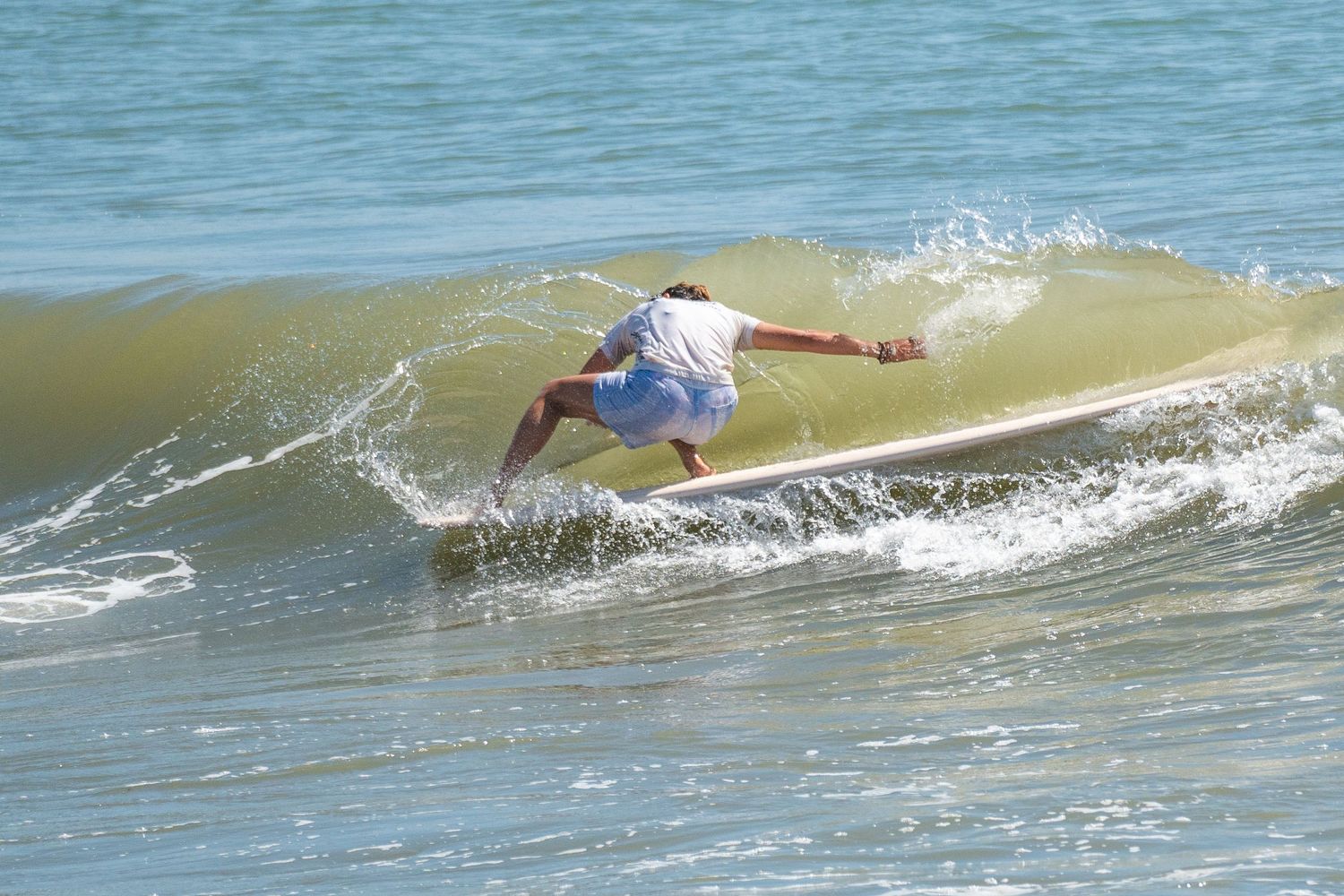 A surfer in a white shirt crouches low on a longboard, carving along the face of a golden-green wave.