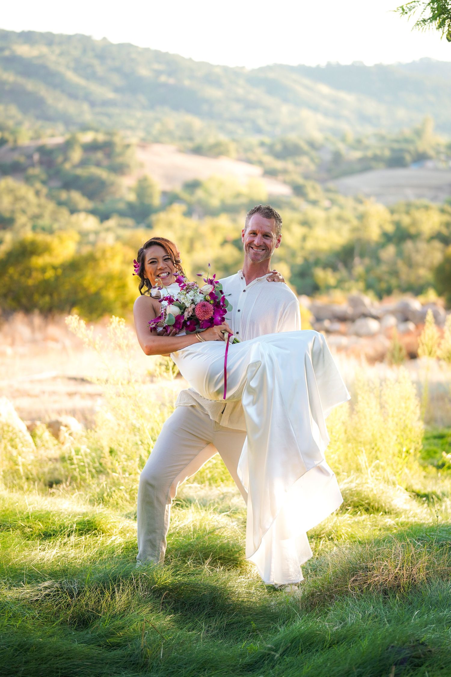 Groom in a white linen shirt lifts his smiling bride in a long white gown, holding her bouquet, on a sunlit grassy hillside.