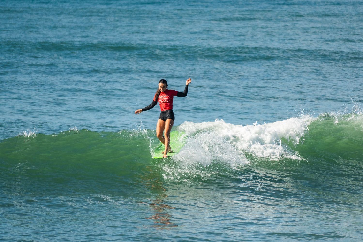 A surfer in a red rash guard balances on a longboard while riding a green wave toward shore.