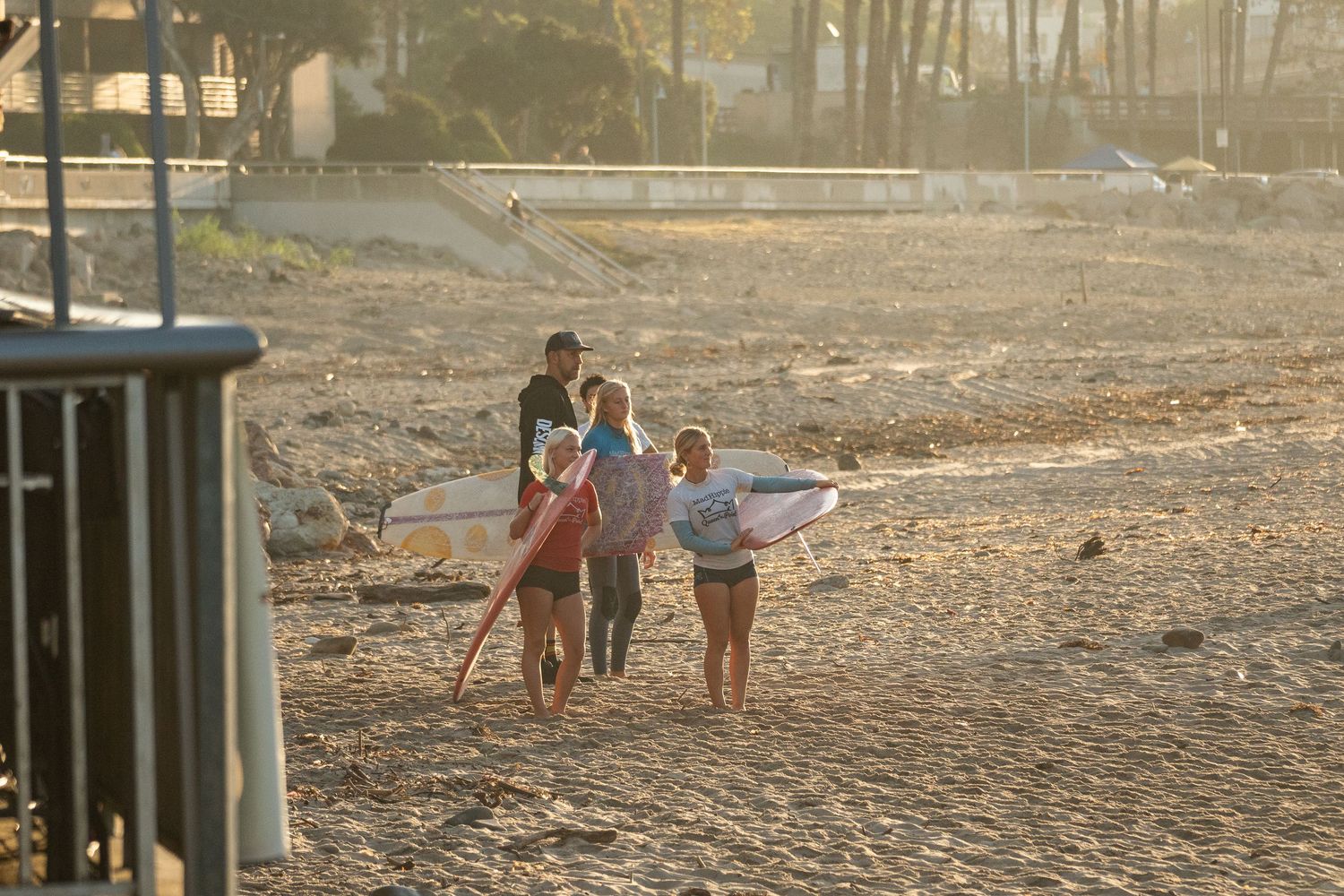 Group of surfers carry their surfboards across the sand toward the water in soft, warm morning light.