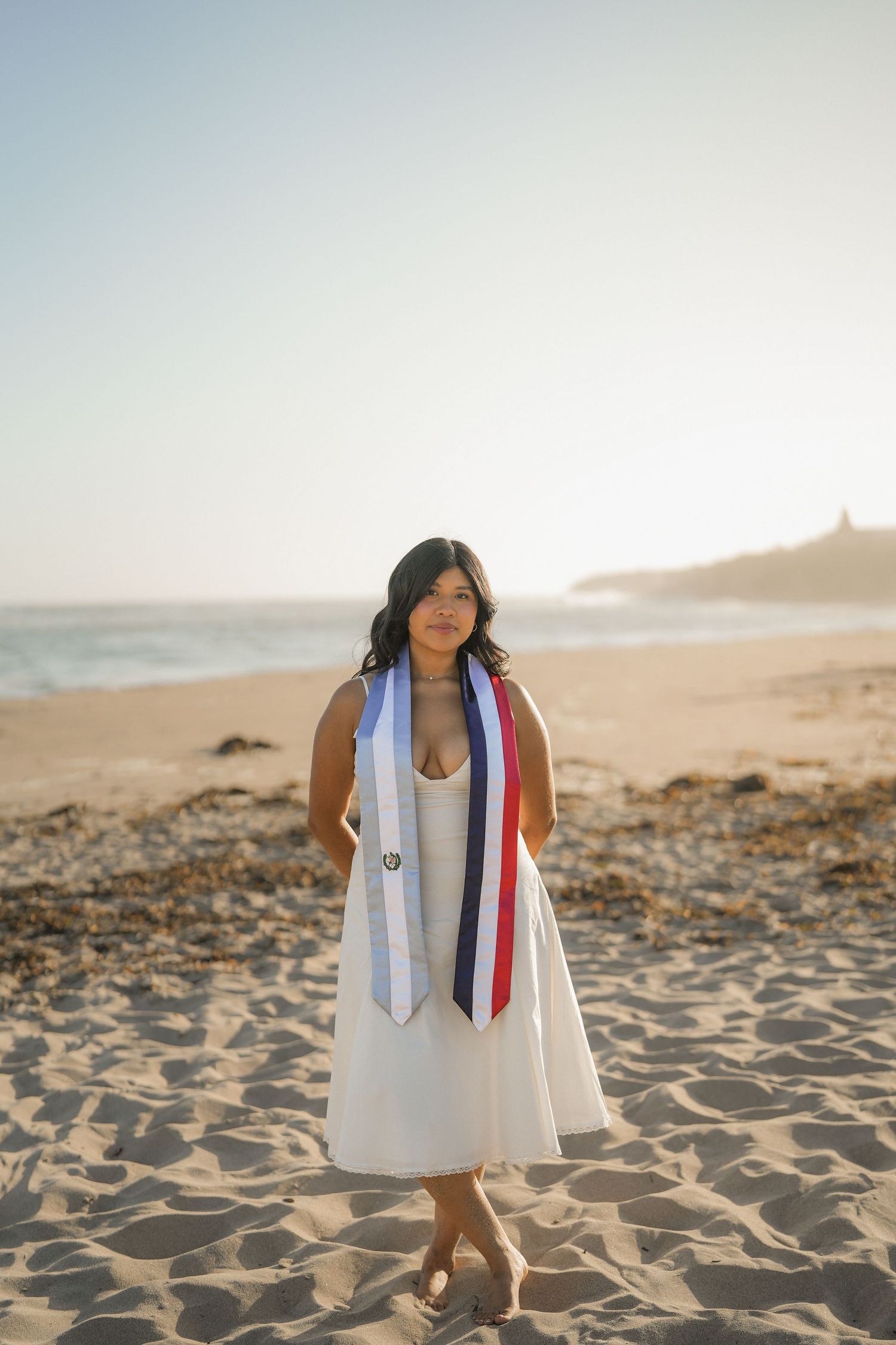 Graduate in a white dress wearing blue, white and red graduation stoles while standing barefoot on a sunny beach.