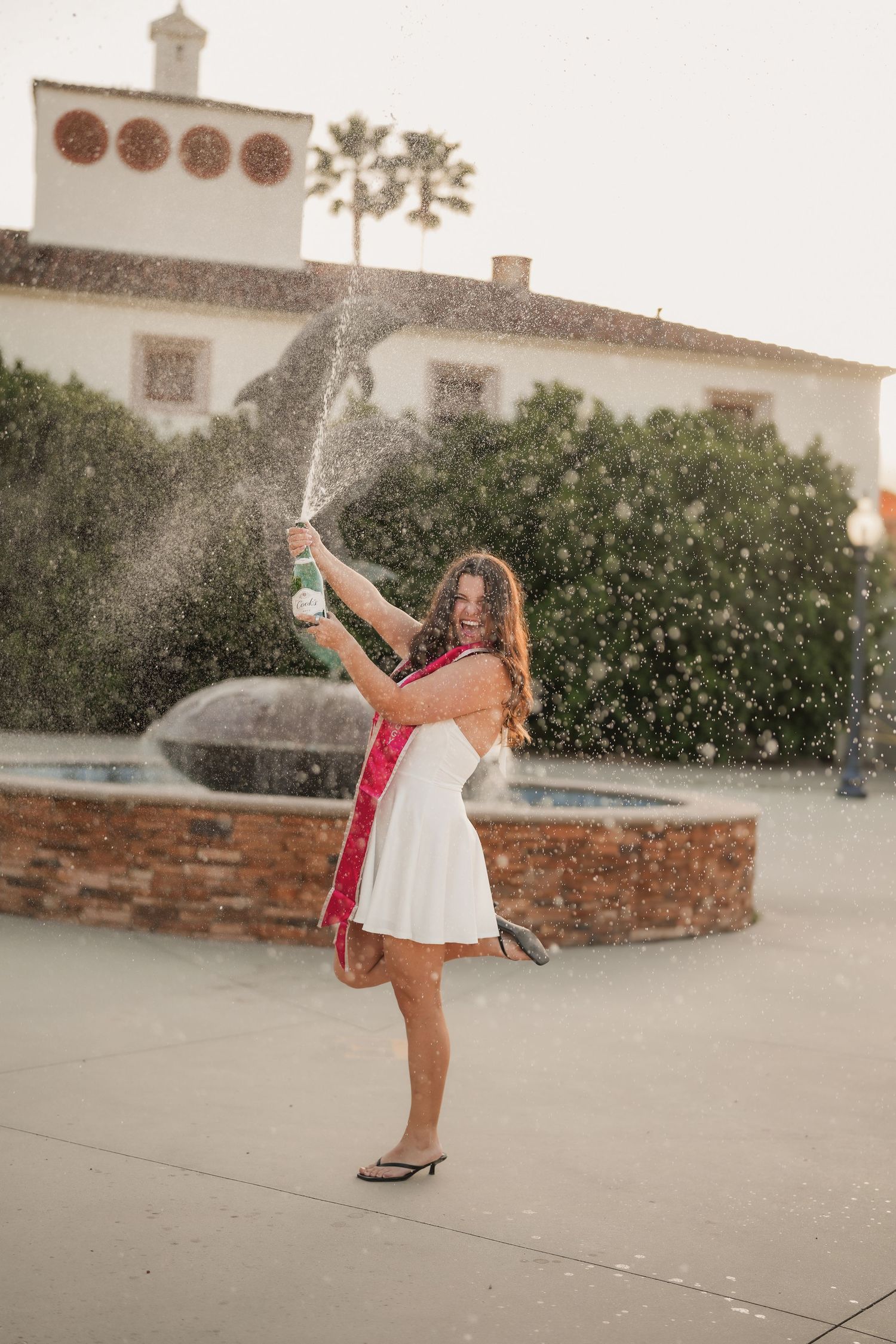 A joyful graduate in a white dress and sash pops a champagne bottle, spraying bubbly beside a fountain and green hedges.