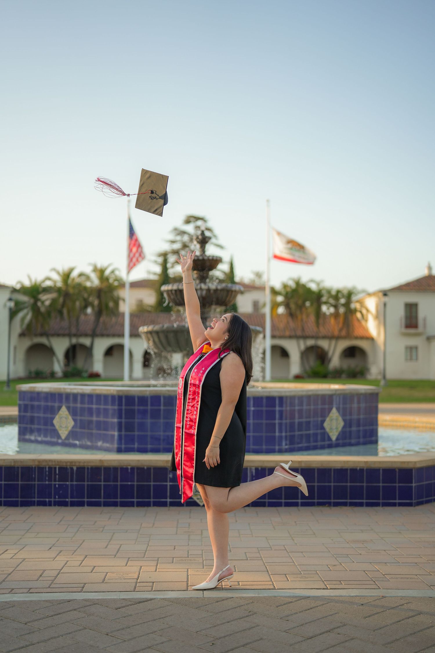 A graduate in a red sash tosses her cap into the air in front of a tiled fountain, palm trees, and the American and California flags.