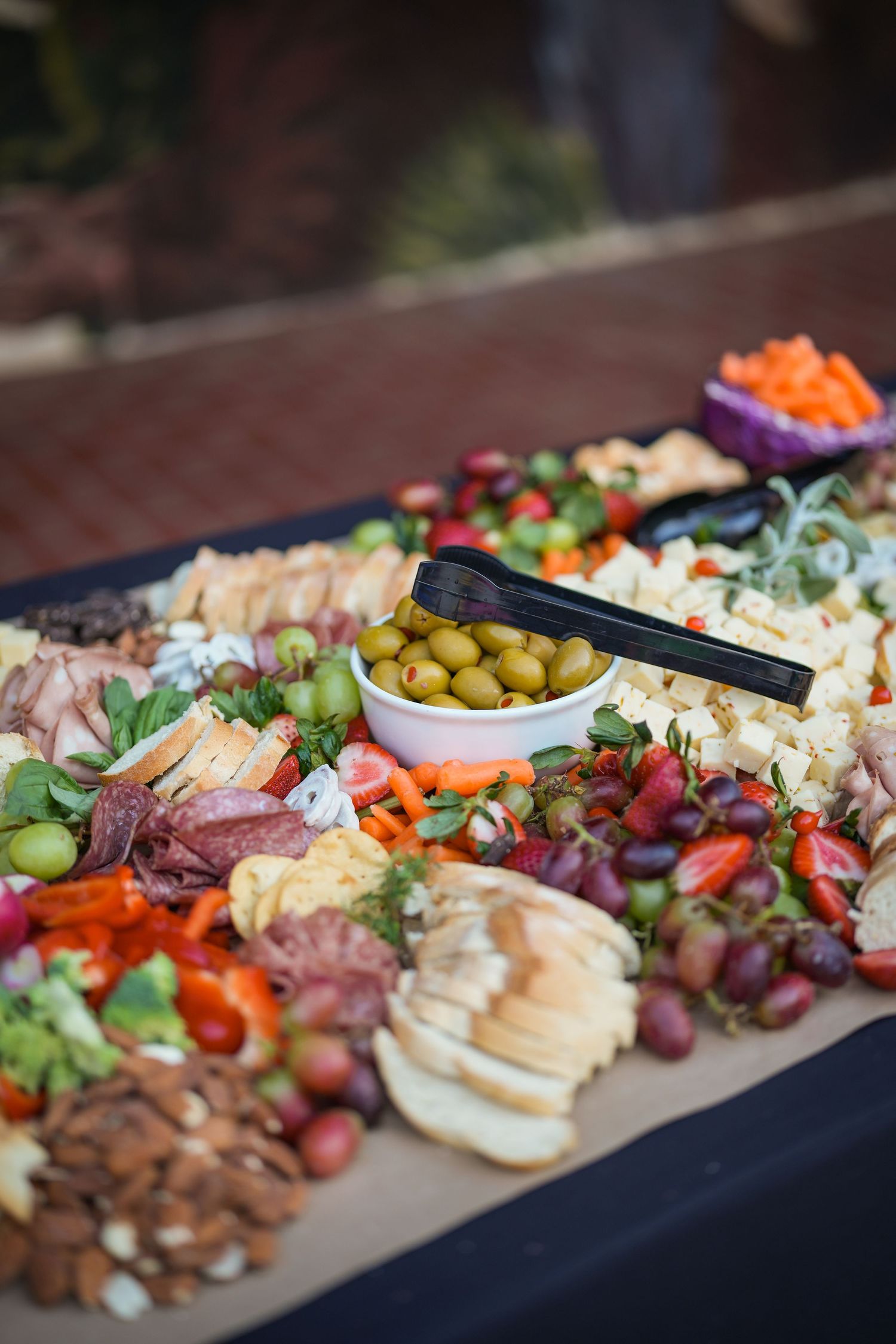 Grazing board with olives, cheeses, cured meats, grapes, sliced bread, peppers and nuts, with black tongs resting on the bowl.