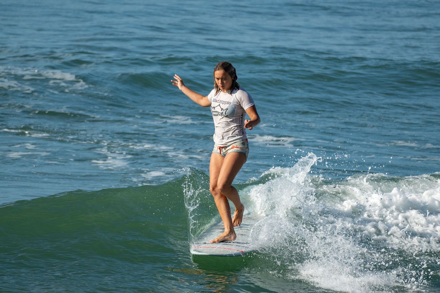 A female surfer in a white shirt rides a longboard on a breaking wave with her arms outstretched.