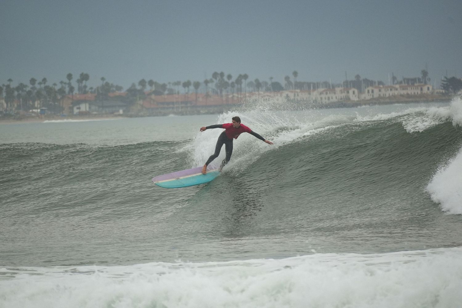 Surfer in a red top carves a turn on a teal surfboard along the face of a large ocean wave.