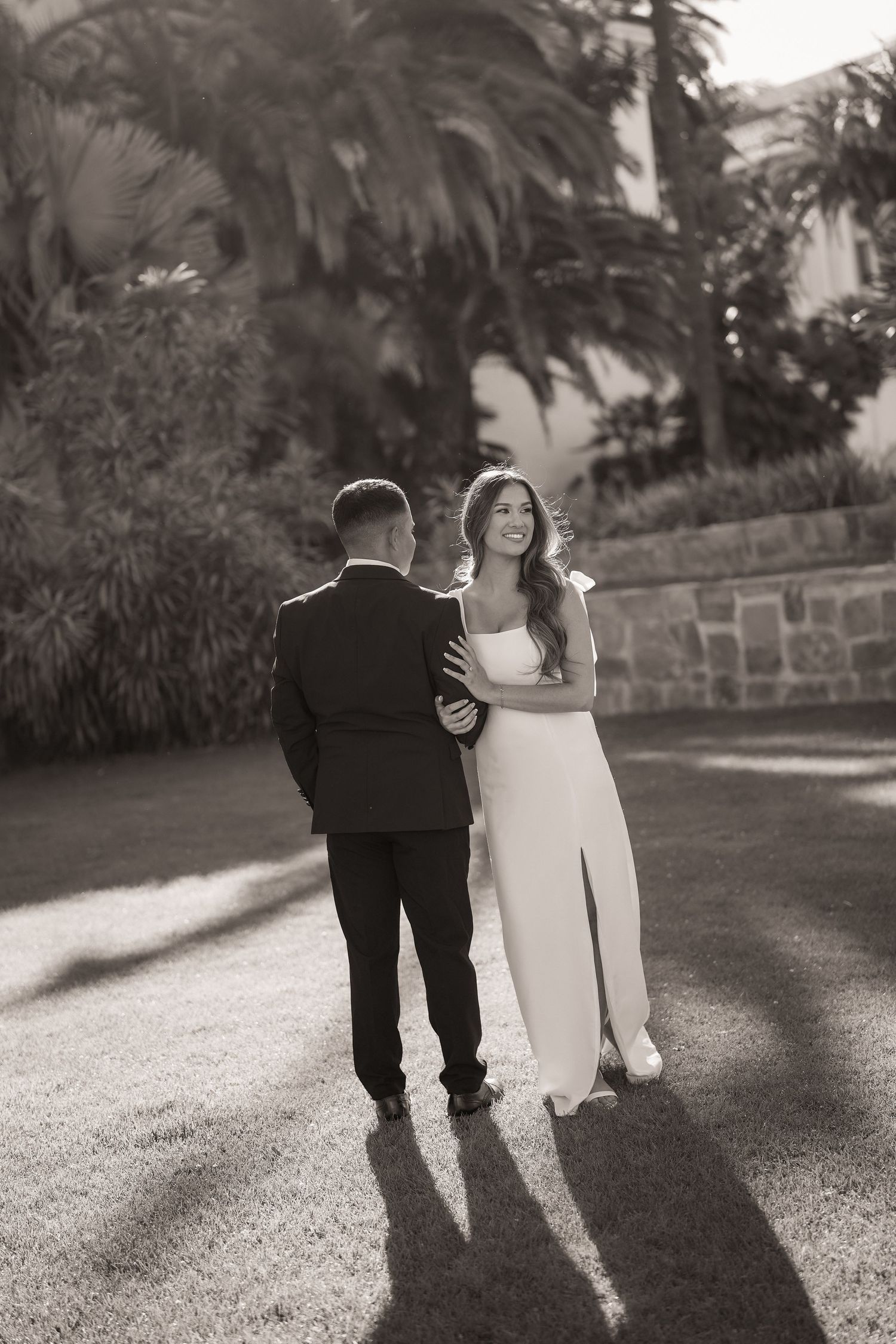 Sepia photo of a bride and groom embracing on a sunlit lawn with palm trees and long shadows.
