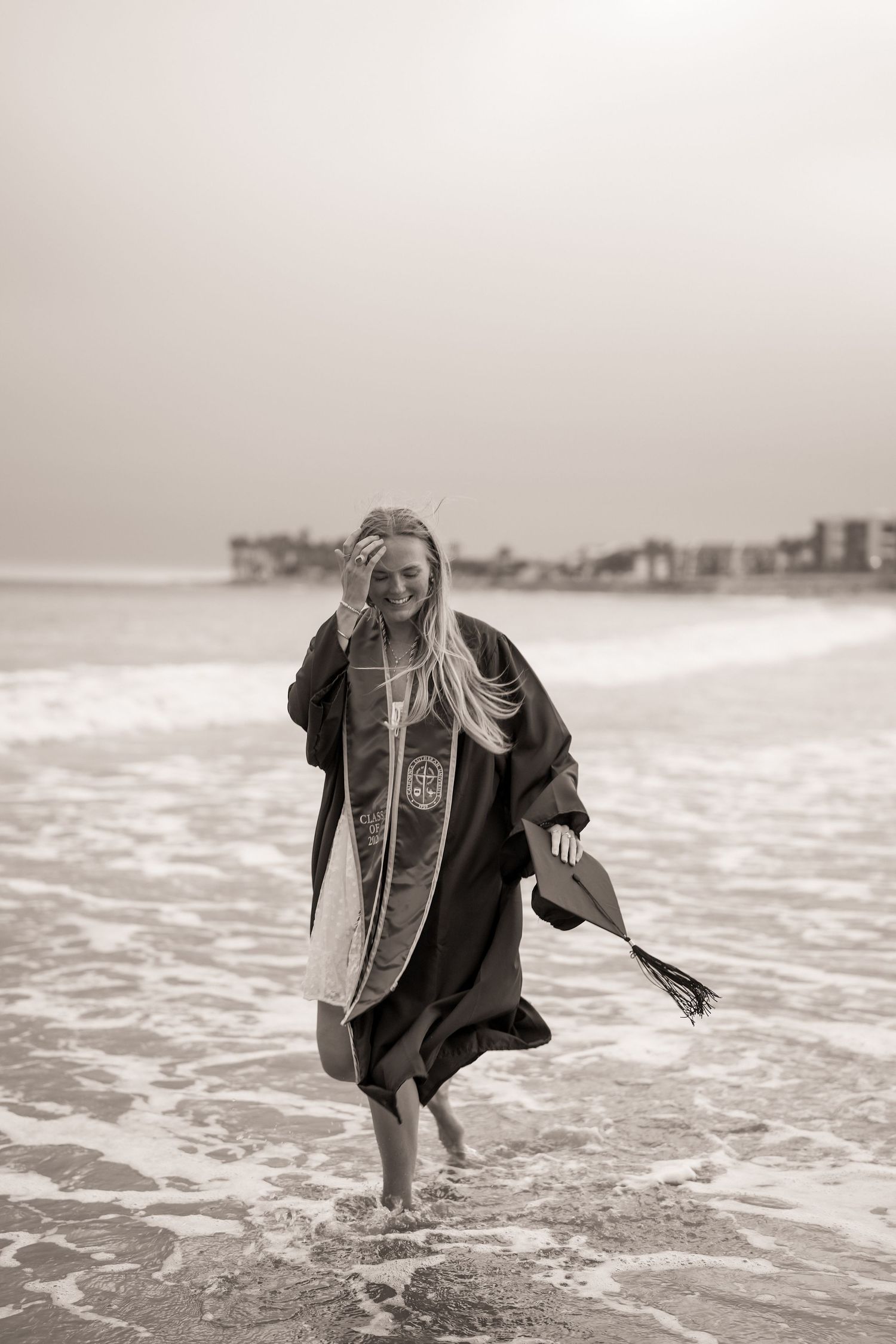 Black and white photo of a graduate in a gown and stole laughing as she walks through shallow ocean waves holding her cap.