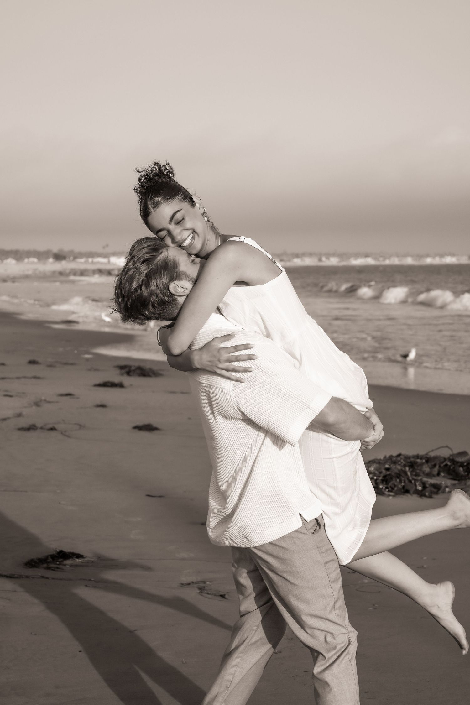 Sepia photo of a joyful man lifting a smiling woman in a white dress in an embrace on a sandy beach by the ocean.