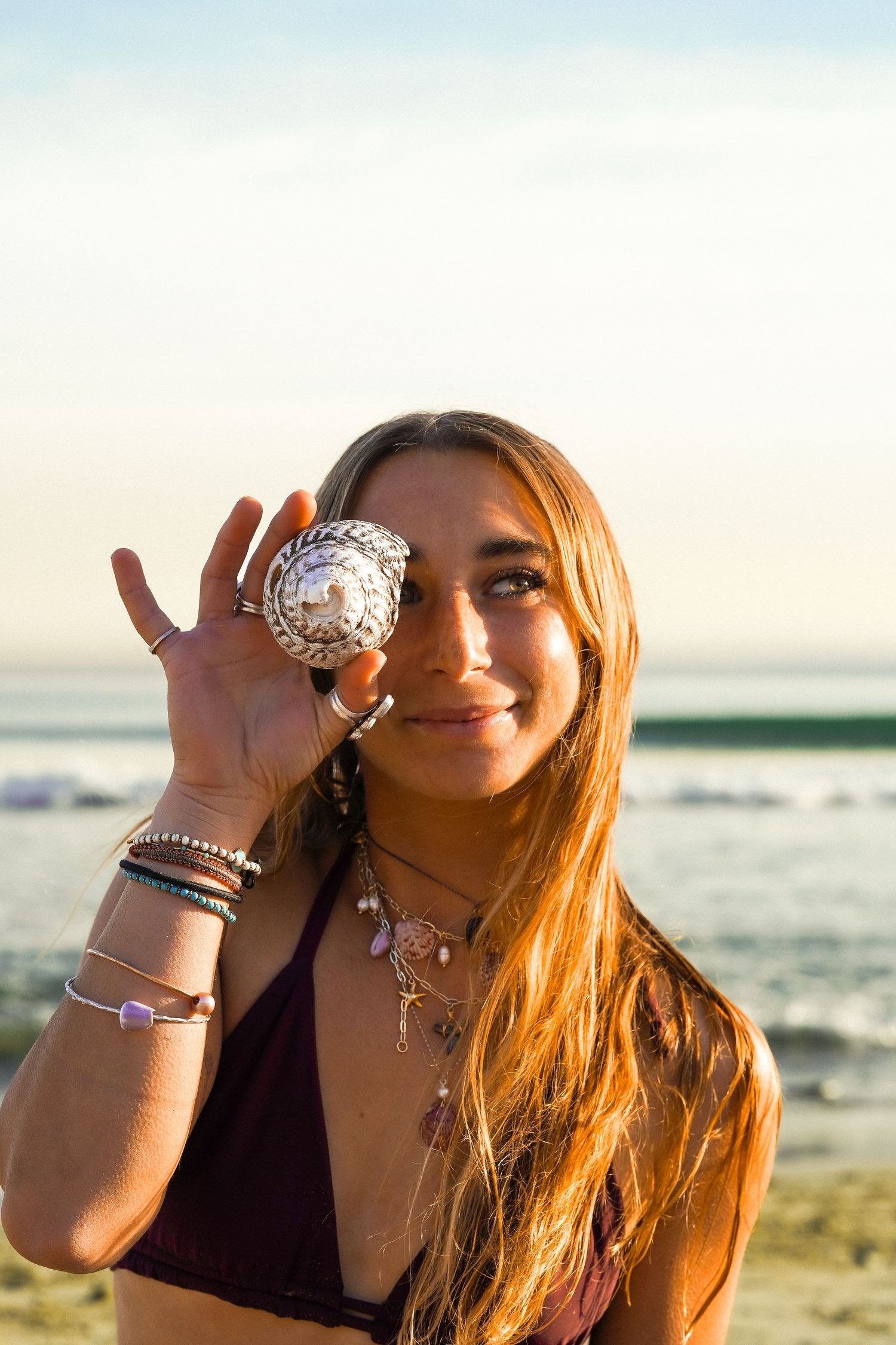 Woman at the beach holds a spiral seashell over one eye, wearing layered necklaces and bracelets with waves behind her.