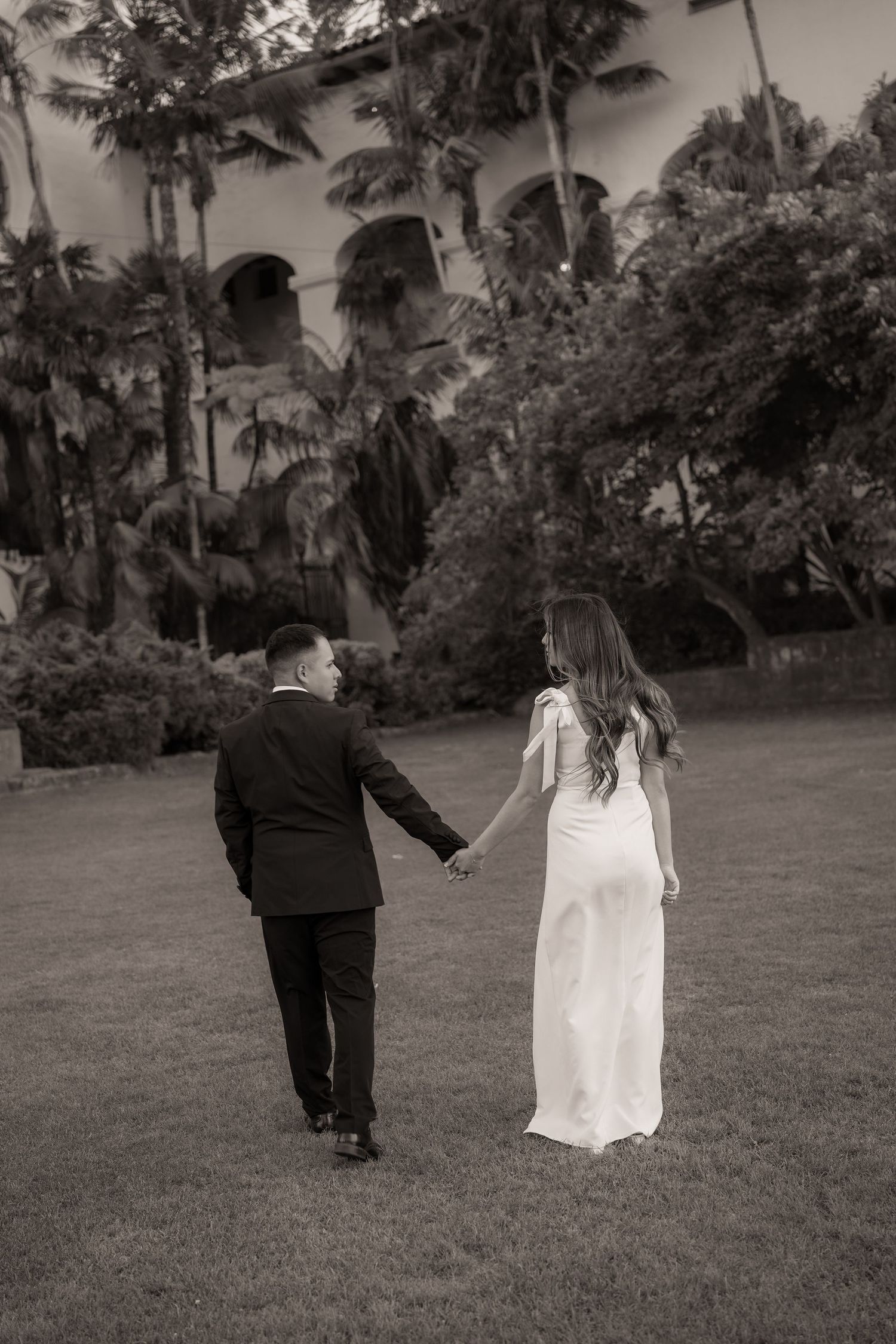 Black and white photo of a bride and groom holding hands and walking across a lawn toward a garden-framed historic arched building.