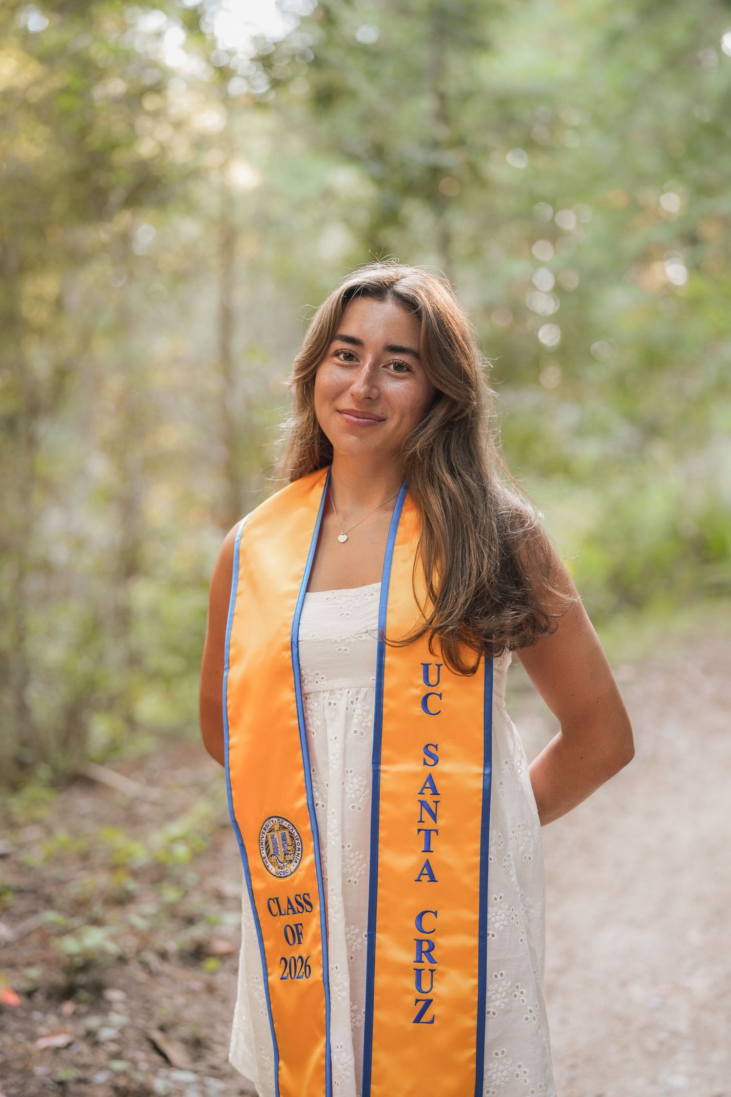 Portrait of a smiling graduate in a gold and blue UC Santa Cruz Class of 2026 stole standing on a sunny forest path.