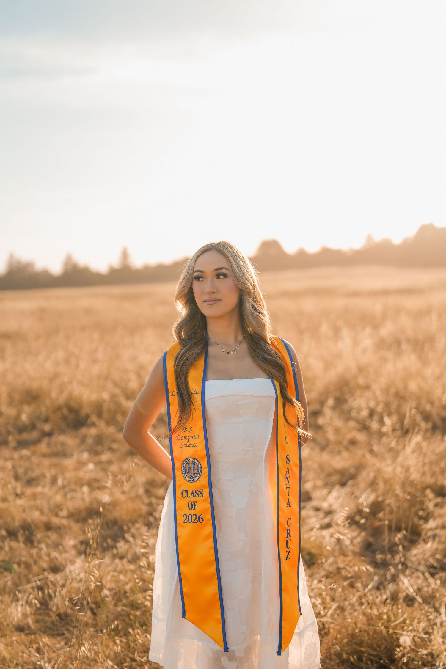 Woman in a white dress wearing an orange Santa Cruz Class of 2026 graduation stole looks aside in a sunlit golden meadow.