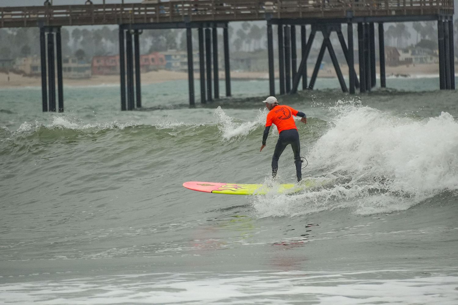 Surfer in an orange shirt rides a pink and yellow longboard on a wave in front of a pier.