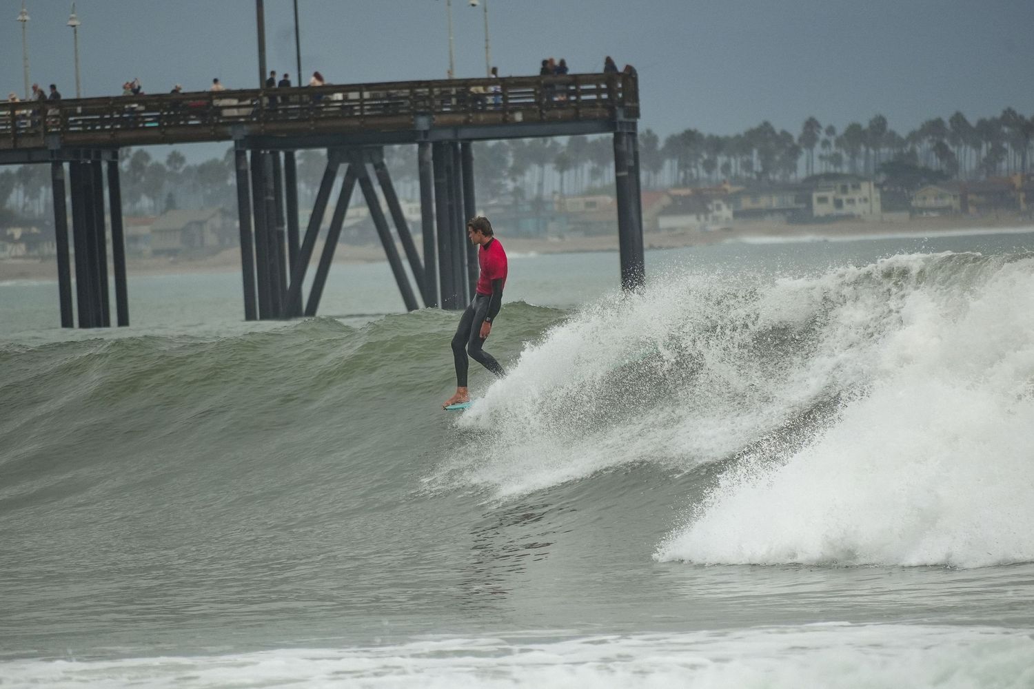 Surfer in a red rash guard rides a curling wave near a wooden pier on an overcast day.