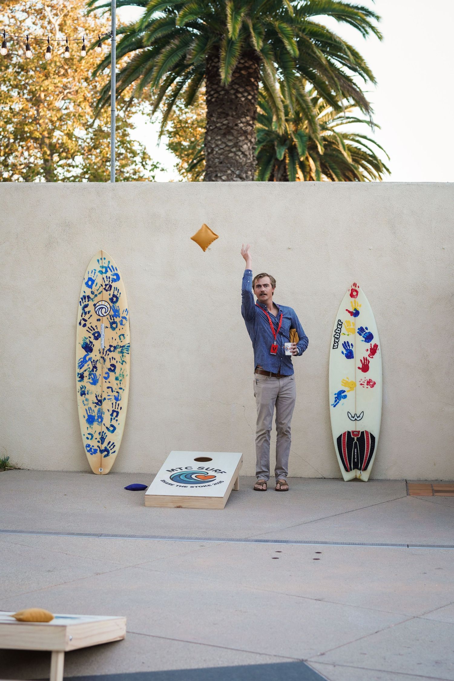 A man tosses a cornhole bag toward a decorated board between two hand-painted surfboards on a sunny outdoor patio.