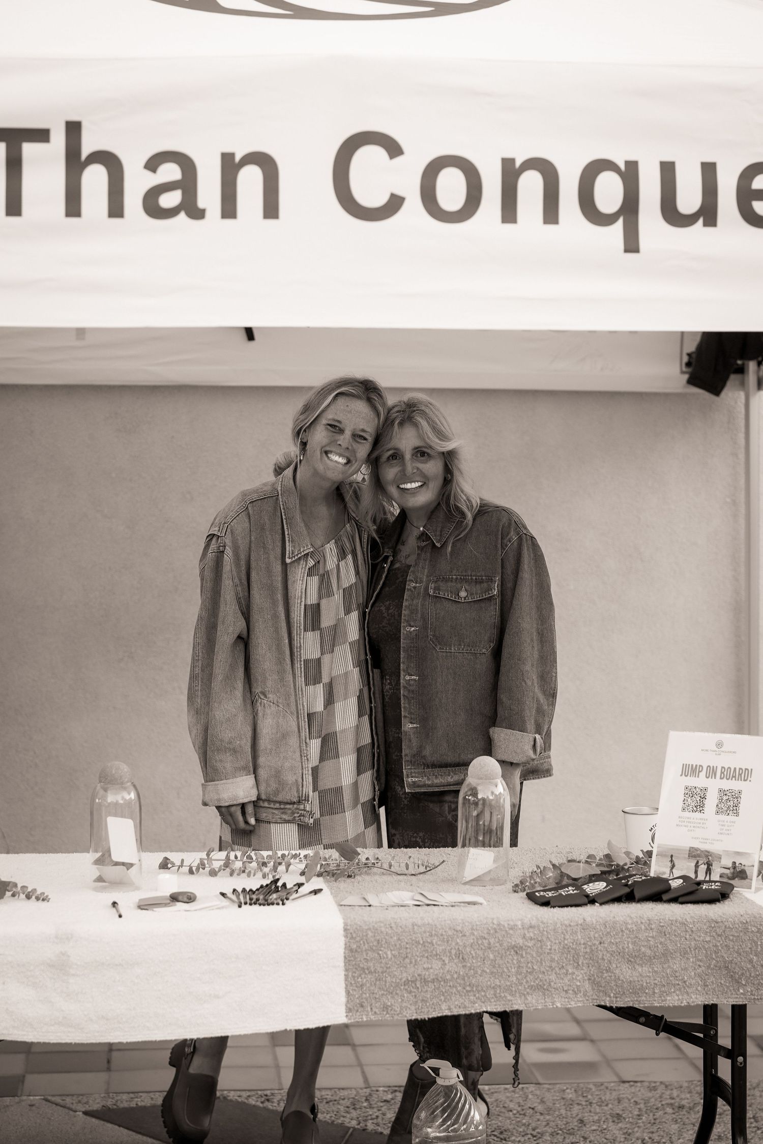 Two smiling women in denim jackets pose behind a vendor table under a More Than Conquerors tent in black and white.