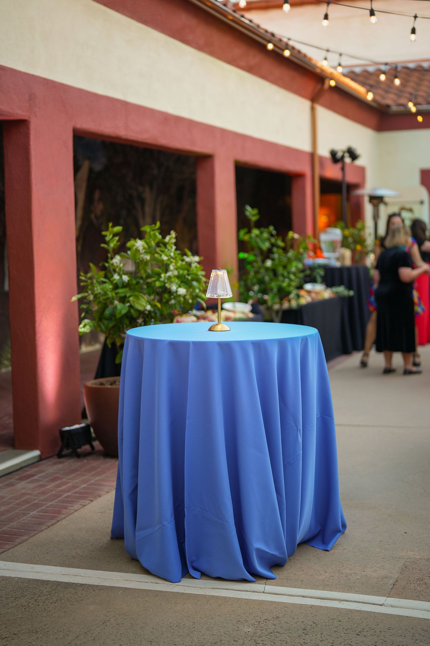 Blue clothed cocktail table with a small gold lamp on a courtyard patio, with guests gathering near buffet tables in the background.