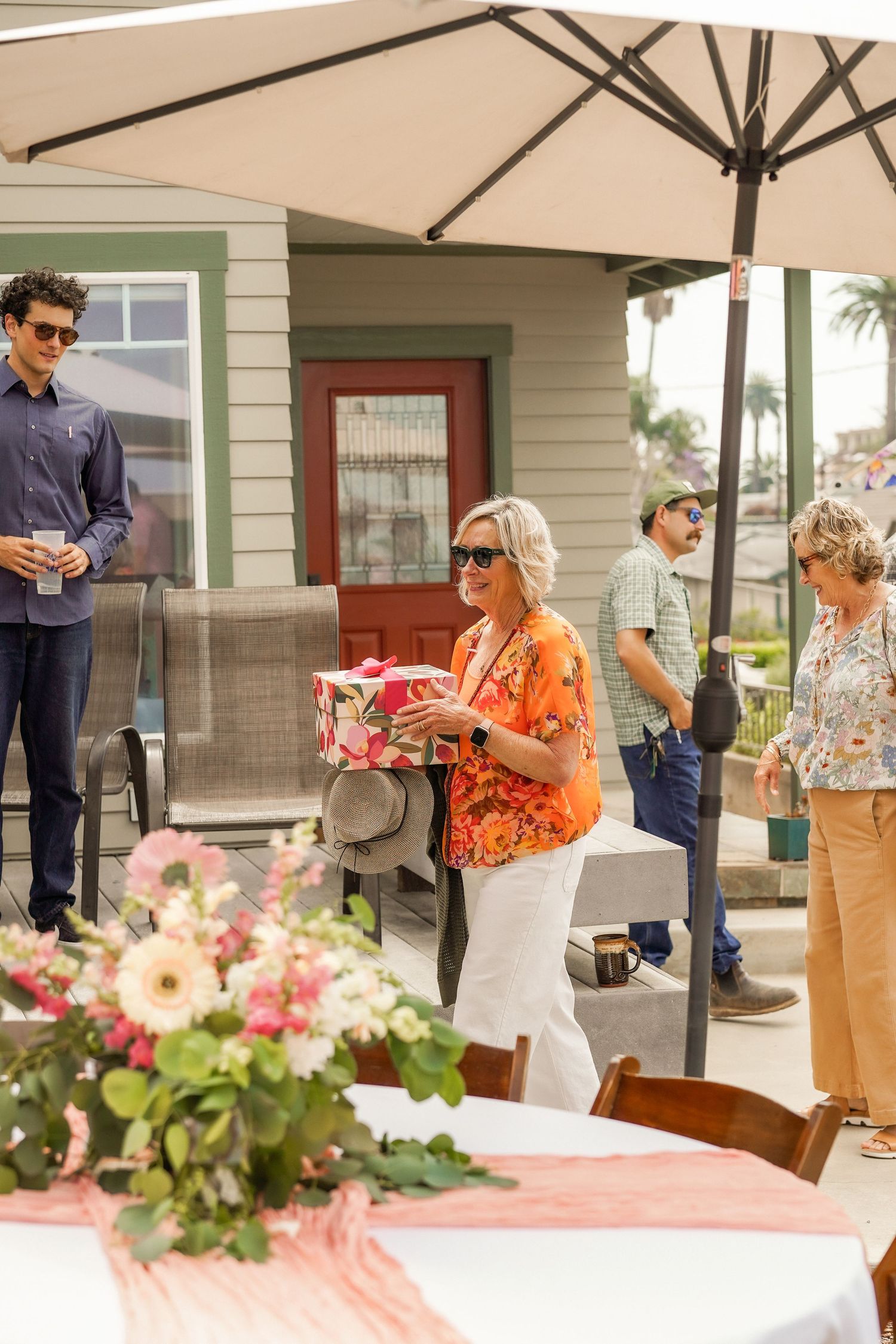 A smiling woman in an orange top holds a pink gift box near a flower arrangement at a patio gathering under an umbrella.