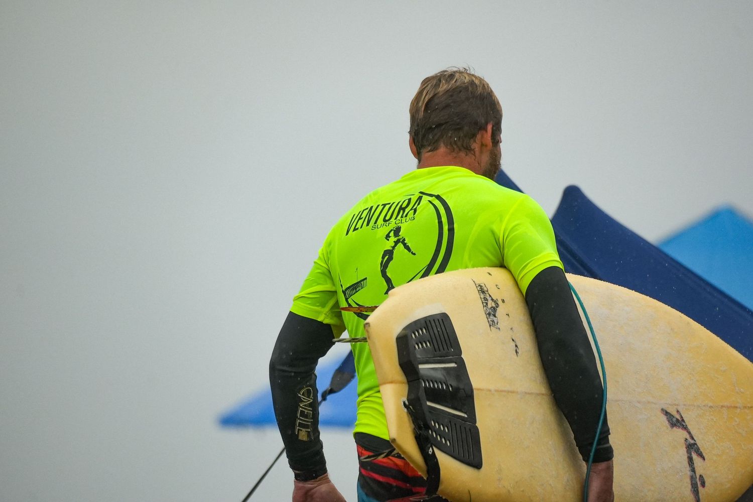 Surfer in a neon green Ventura shirt carries a yellow surfboard under his arm on an overcast beach.