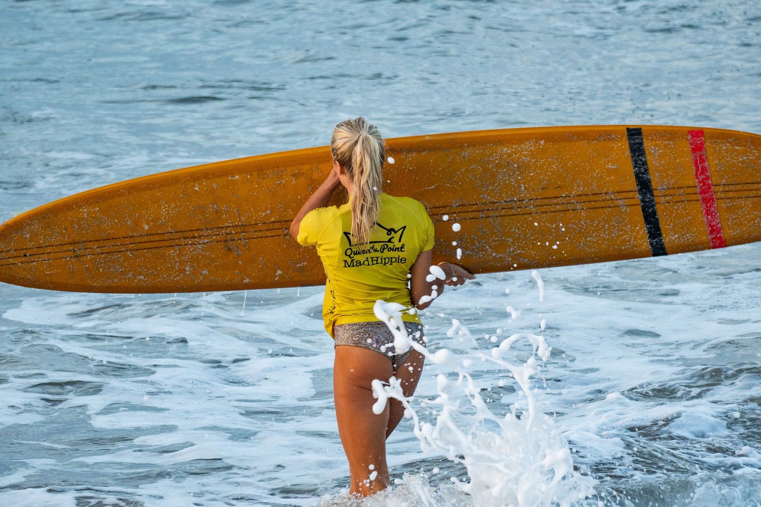 Blonde woman in a yellow shirt carries an orange longboard with black and red stripes while ocean water splashes around her.