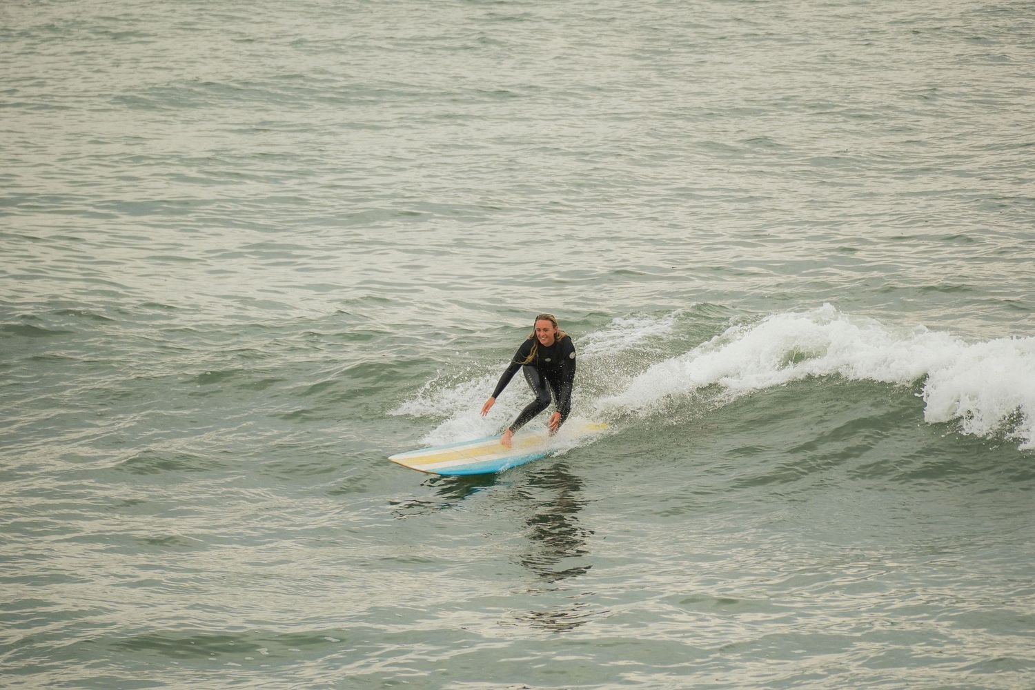 Surfer in a wetsuit crouches low on a colorful surfboard while riding a breaking wave in calm gray water.