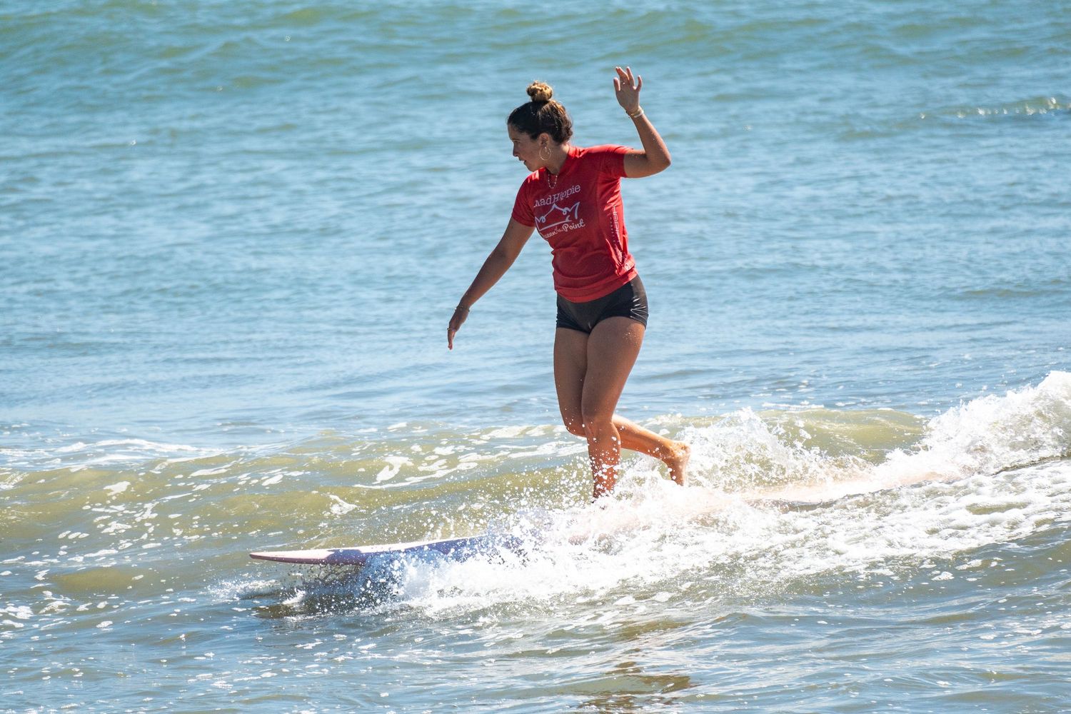 Female surfer in a red shirt balances on a longboard with one hand raised while riding whitewater in the ocean.