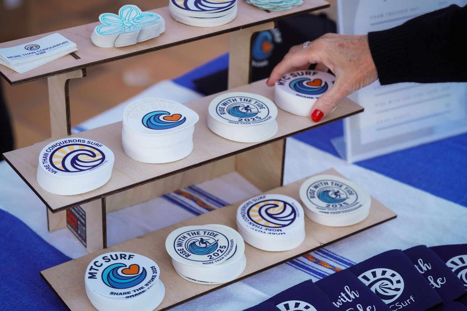 Hand reaches for round MTC Surf wave logo stickers displayed on tiered wooden shelves at an event table.