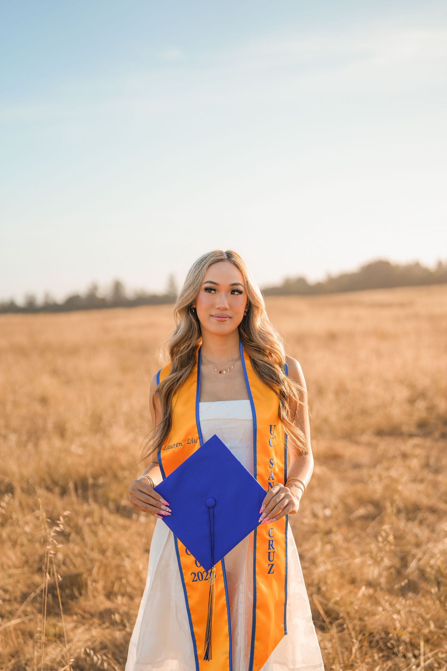 Graduate in an orange UC Santa Cruz stole holds a blue graduation cap while standing in a golden field at sunset.