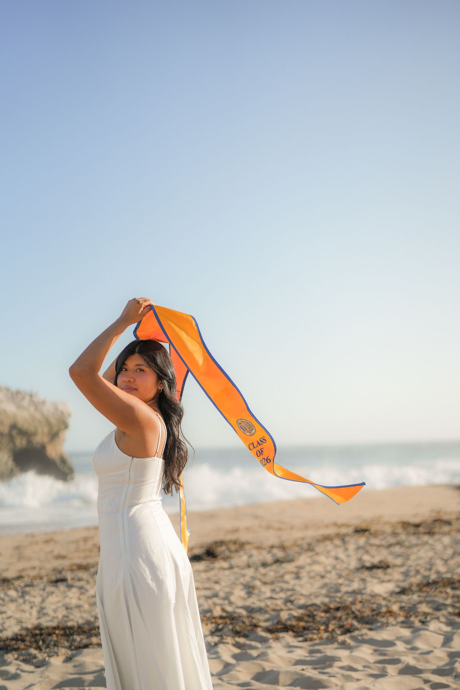 Woman in a white dress holding an orange graduation sash above her head on a breezy beach with crashing waves.
