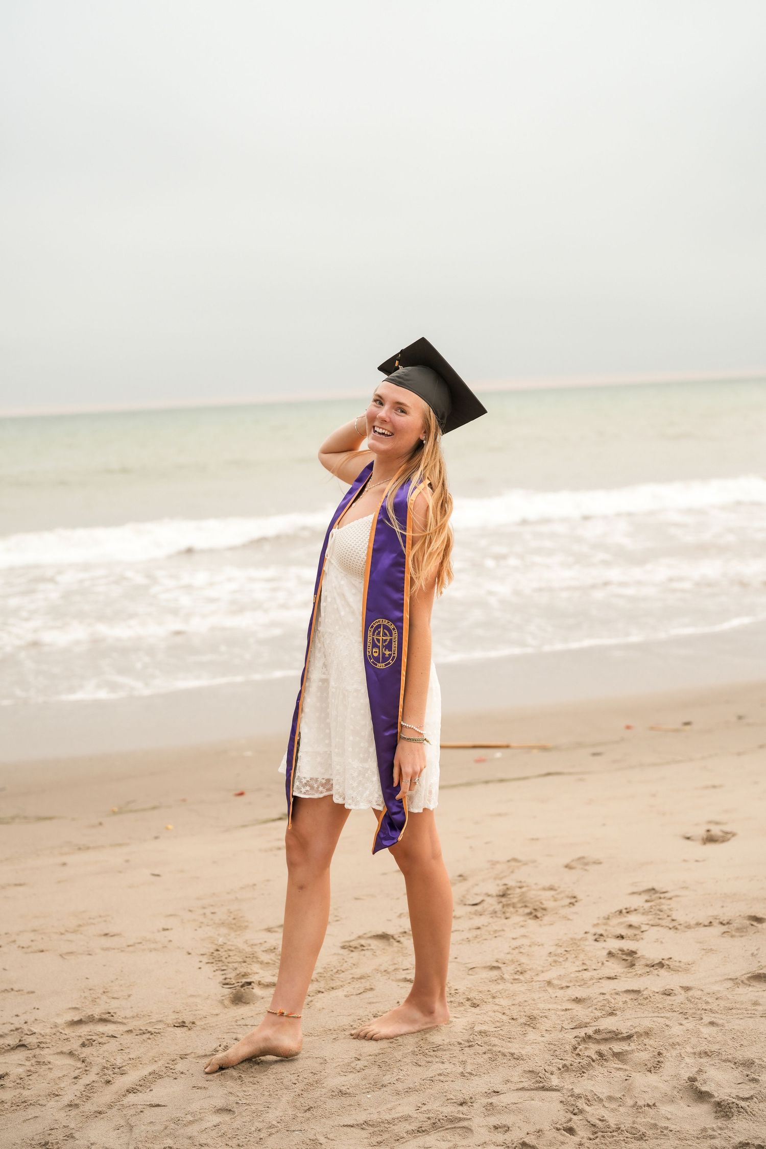 Smiling graduate in a cap, white lace dress and purple stole poses barefoot on a sandy beach by the ocean.