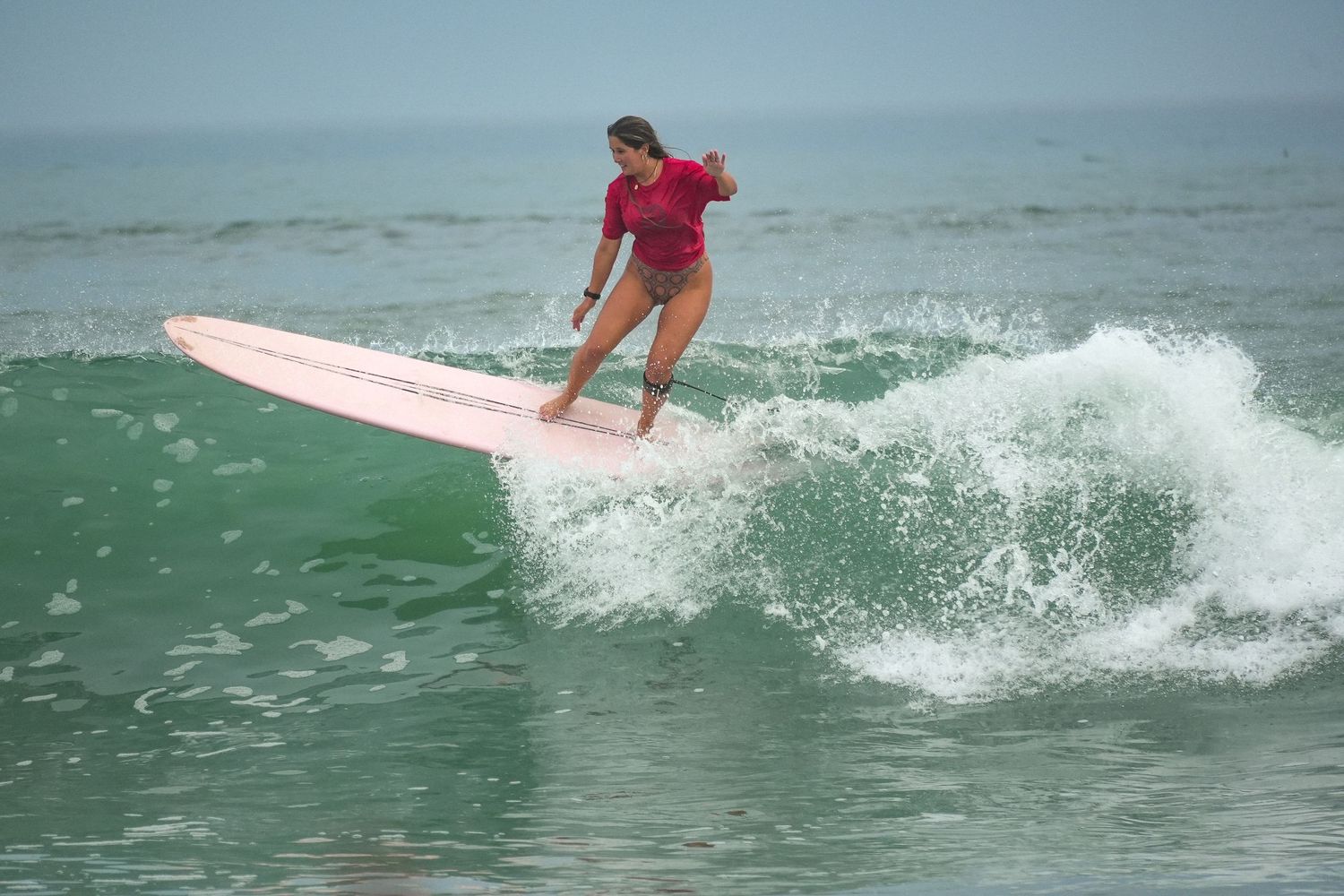 Female surfer in a red top rides a long pink longboard across a small green wave with white foam.