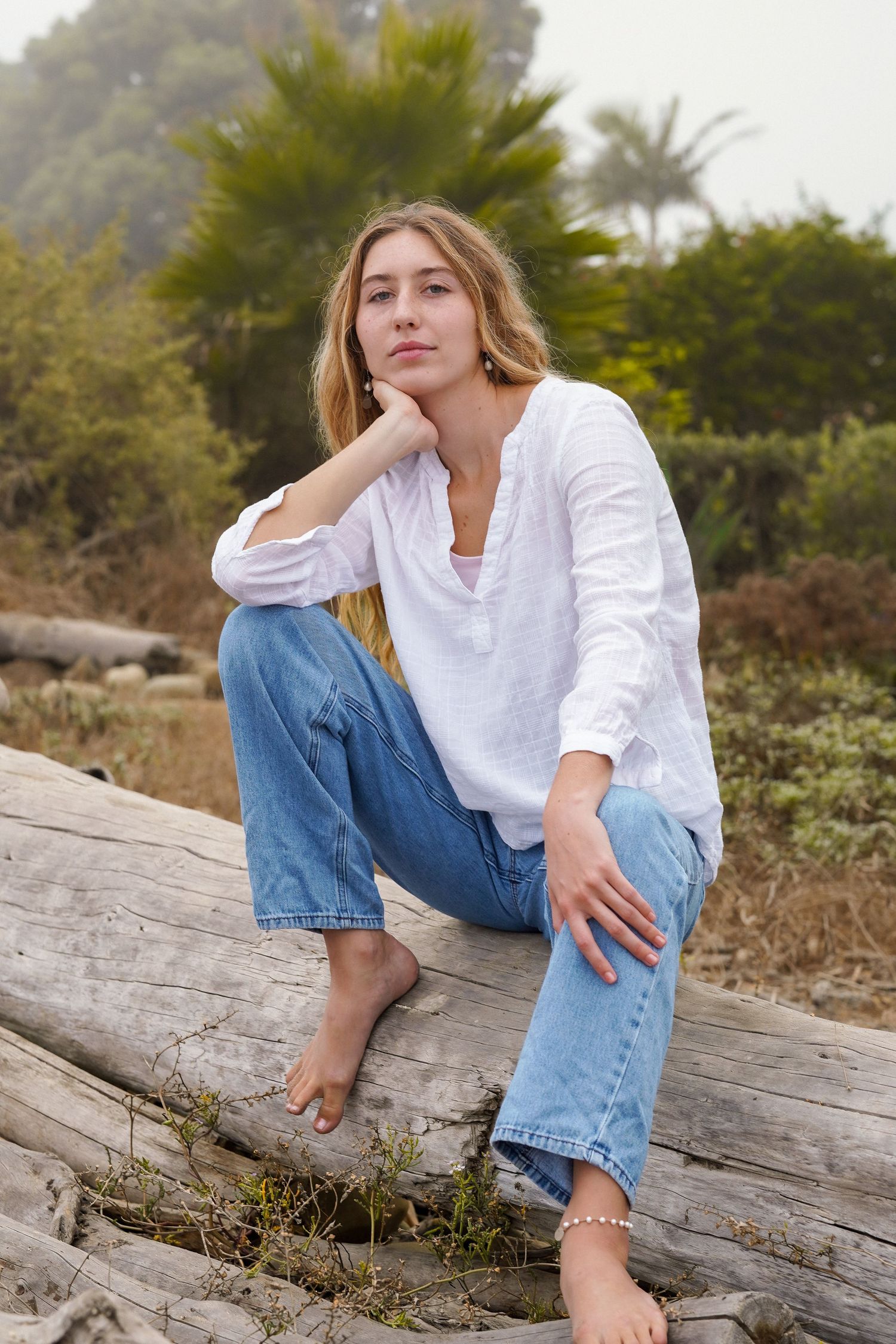 Woman in a white linen shirt and blue jeans sits barefoot on a weathered driftwood log with her hand resting on her chin.