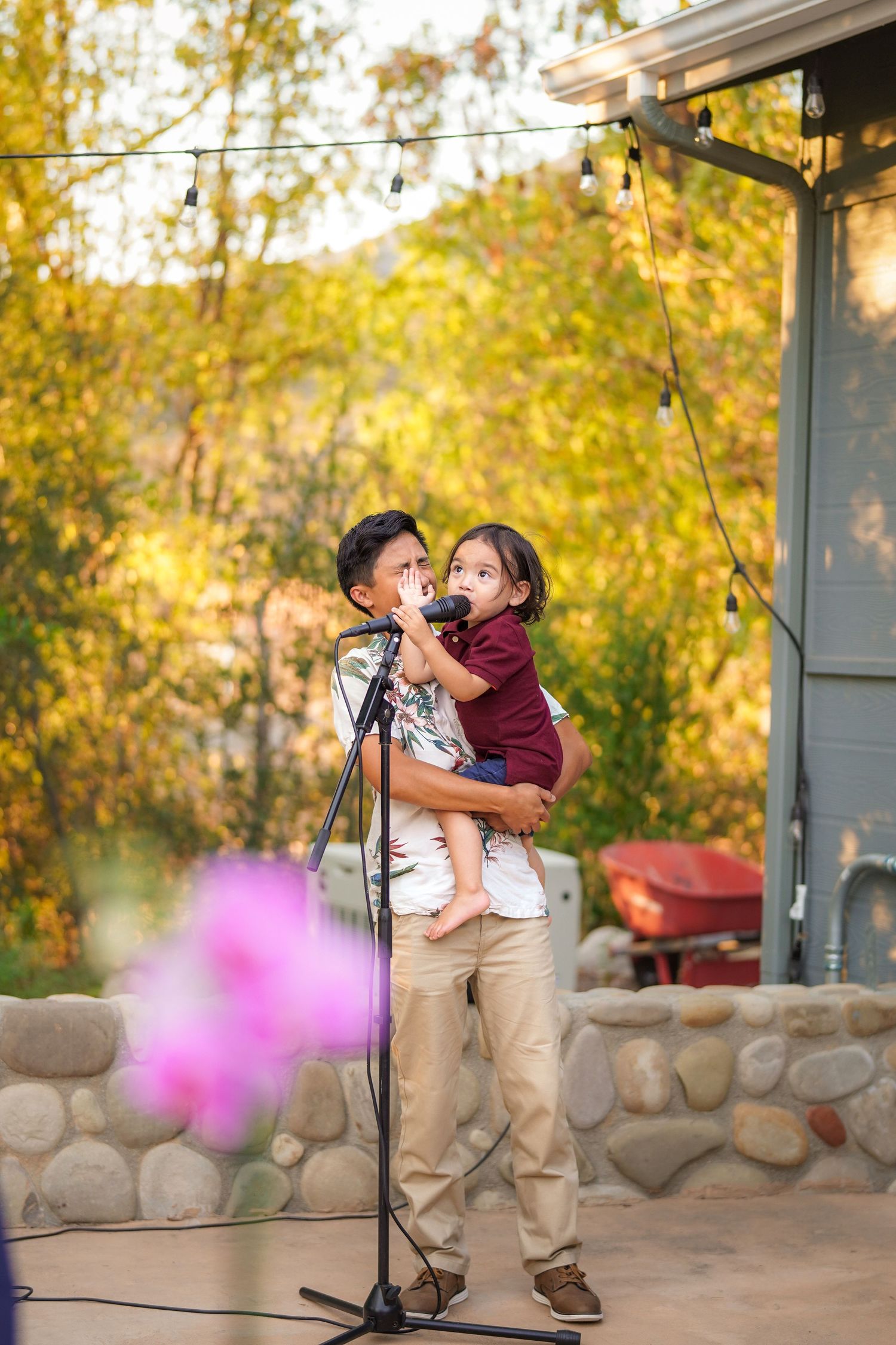Man holds a small child in his arms as the child speaks into a microphone at an outdoor event with string lights and autumn trees.