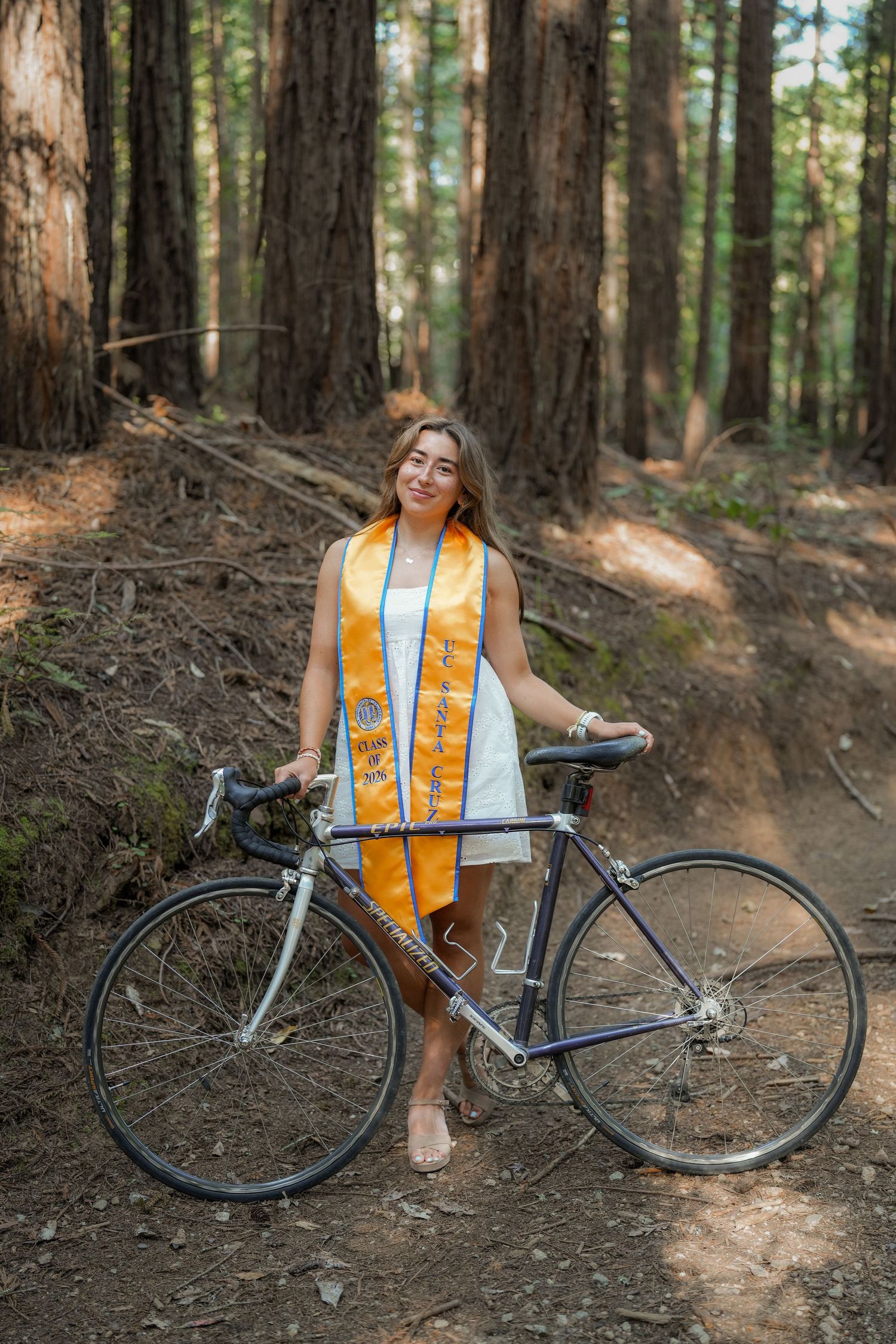 Smiling graduate in a gold stole and white dress leans on a vintage bicycle on a redwood forest trail.