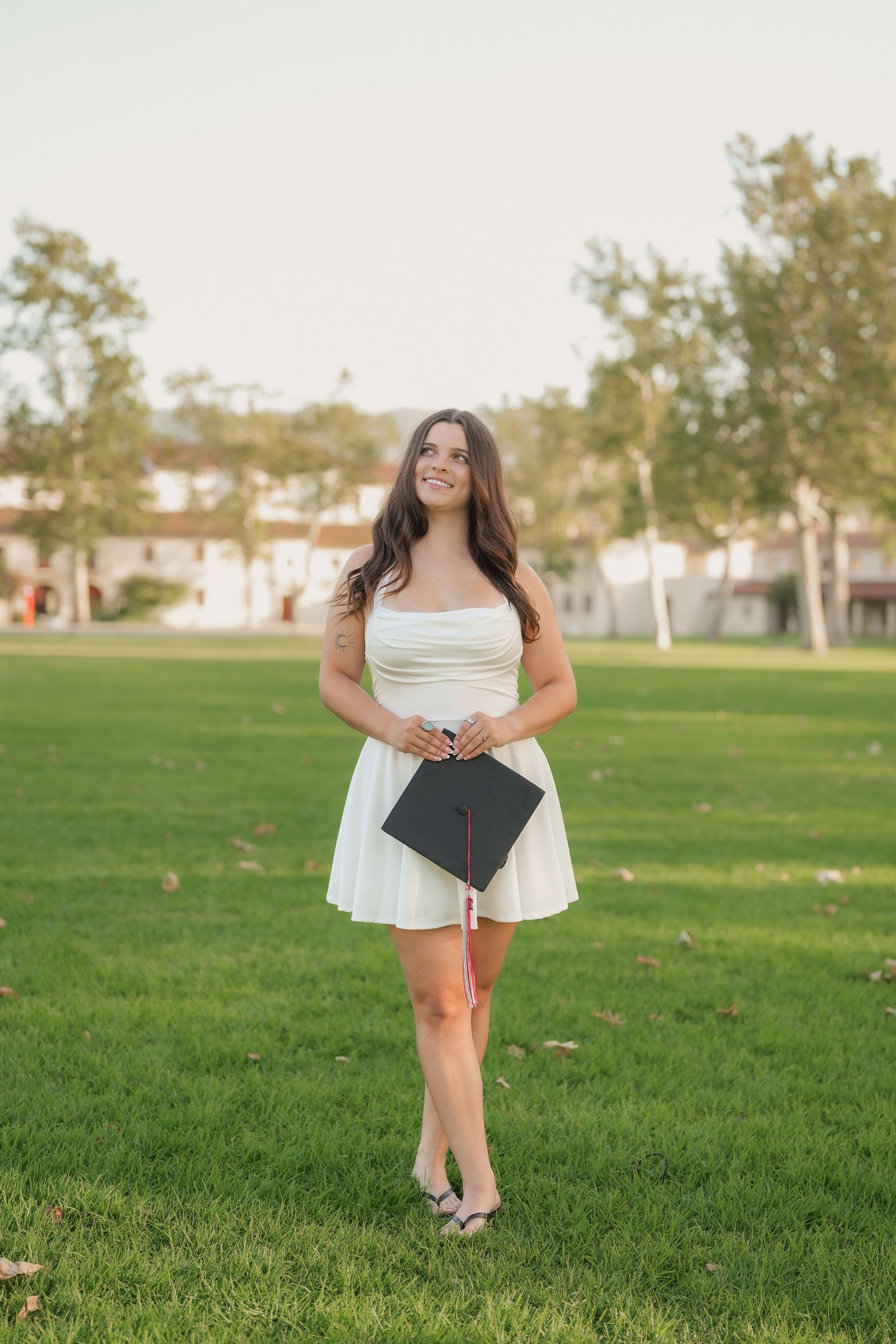 Graduate in a white dress holds a black graduation cap with a pink tassel while standing on a green lawn with trees behind.