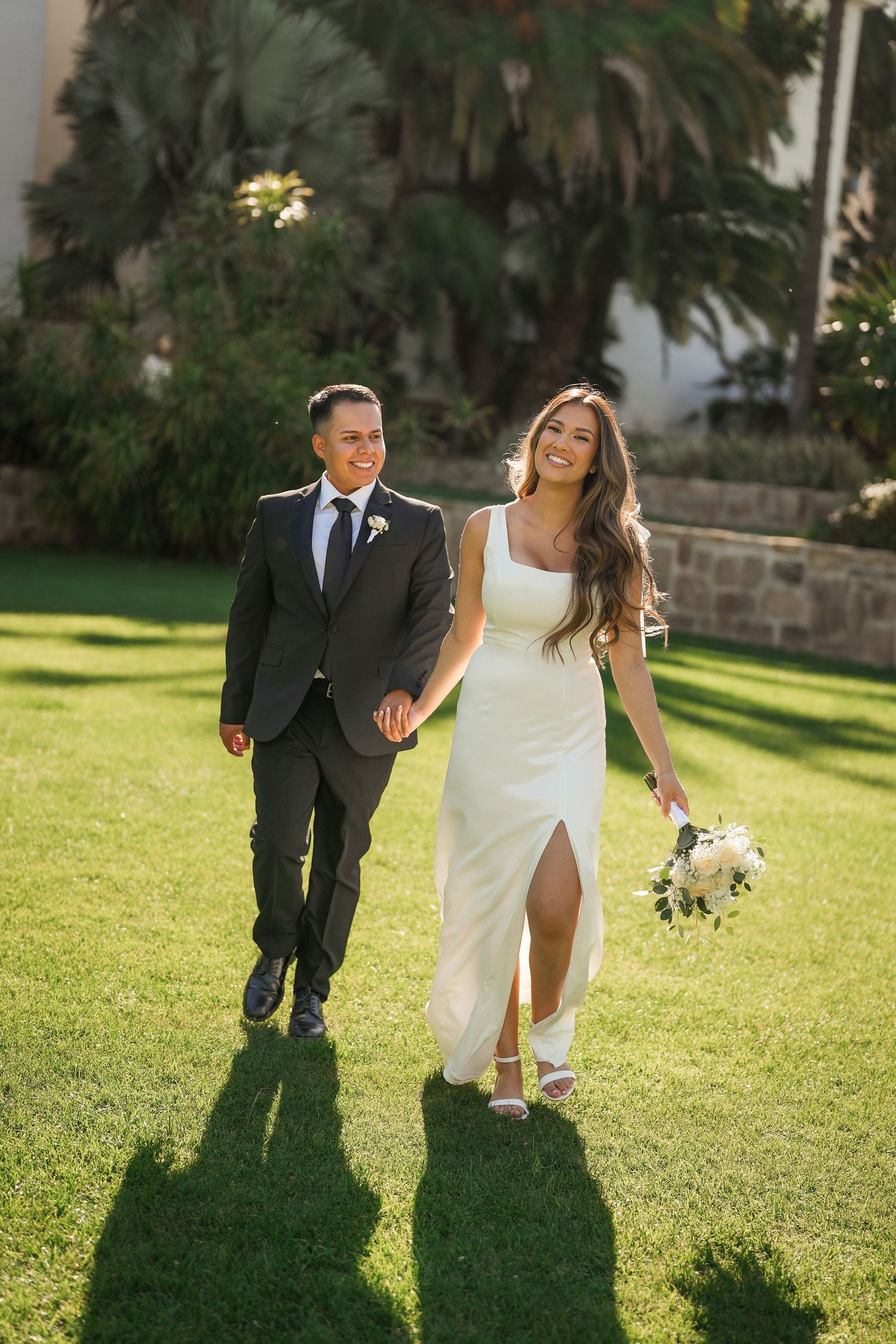 Smiling bride in a white slit gown holding a bouquet walks hand in hand with her groom across a sunny green lawn.