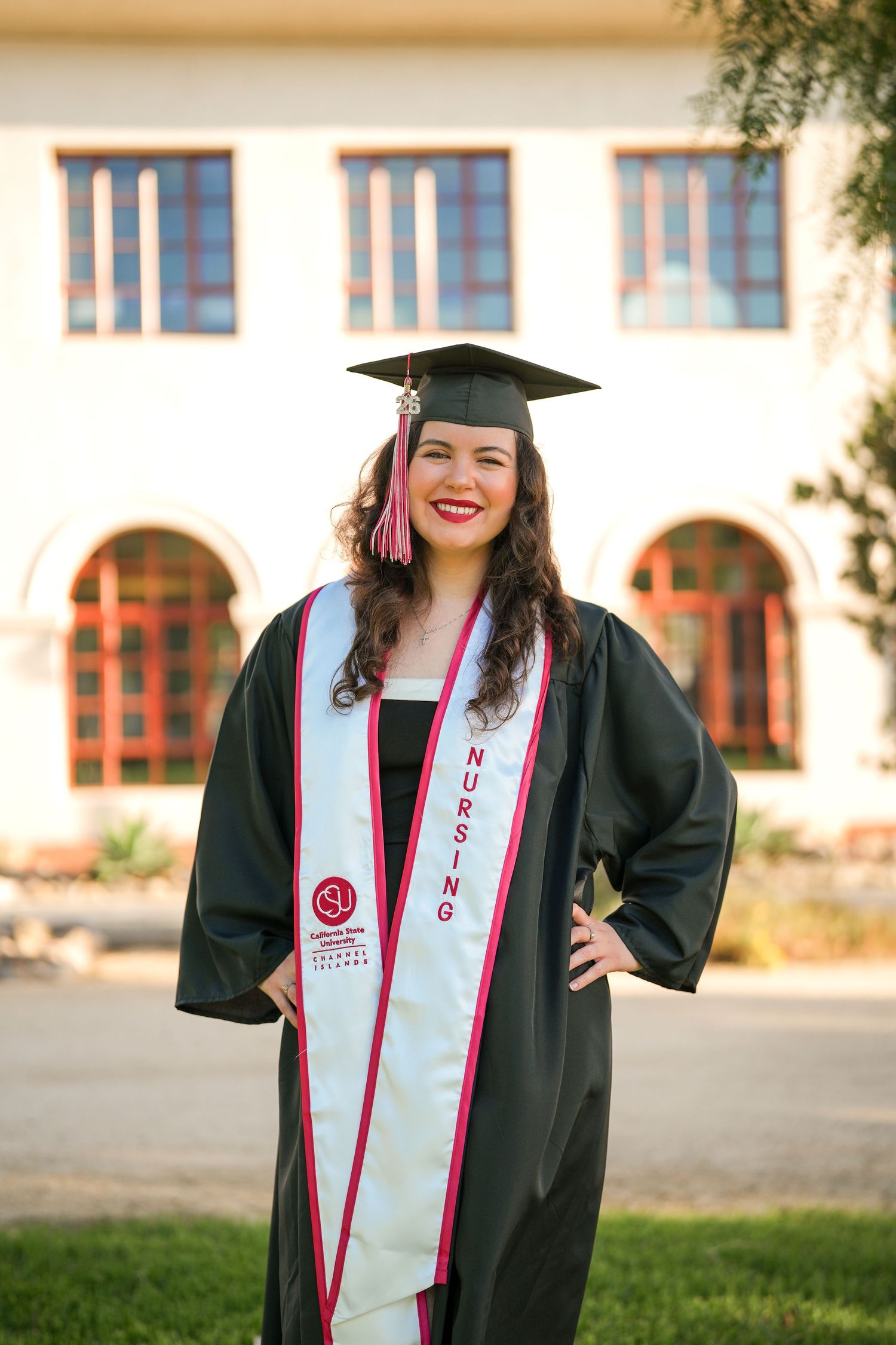 A smiling nursing graduate in a black gown, cap with red tassel, and white nursing stole poses with hands on hips outside a building.