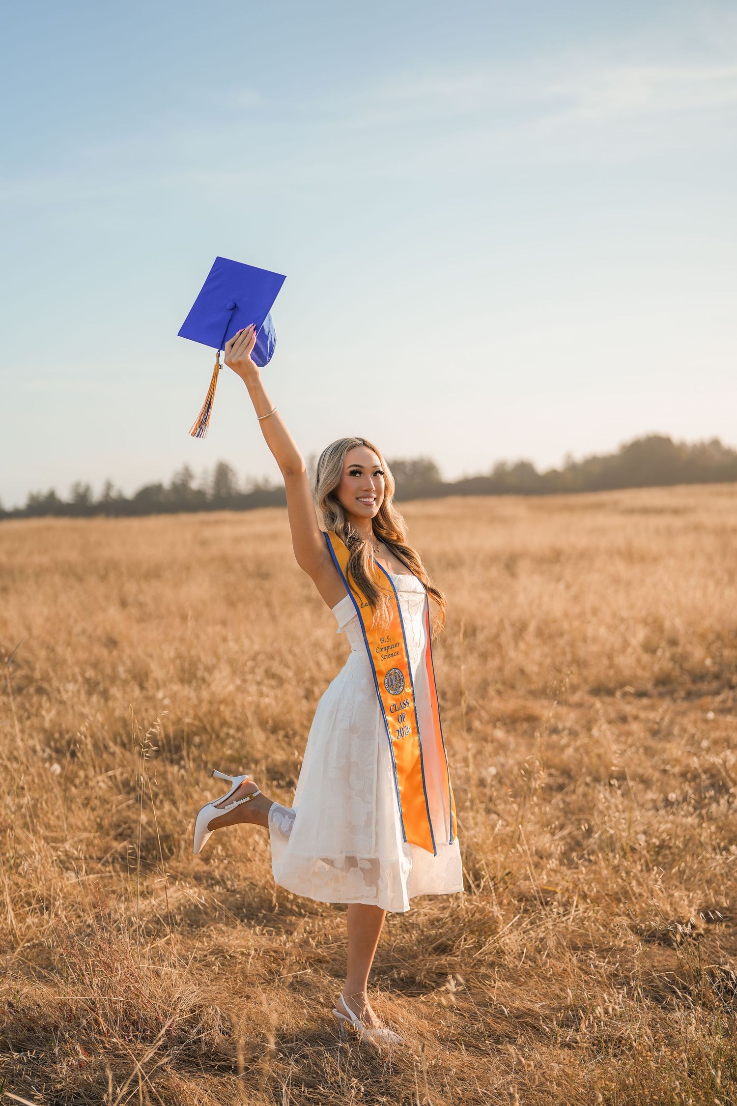 Joyful graduate in a white dress and orange stole kicks up a heel and raises a blue cap in a golden field.