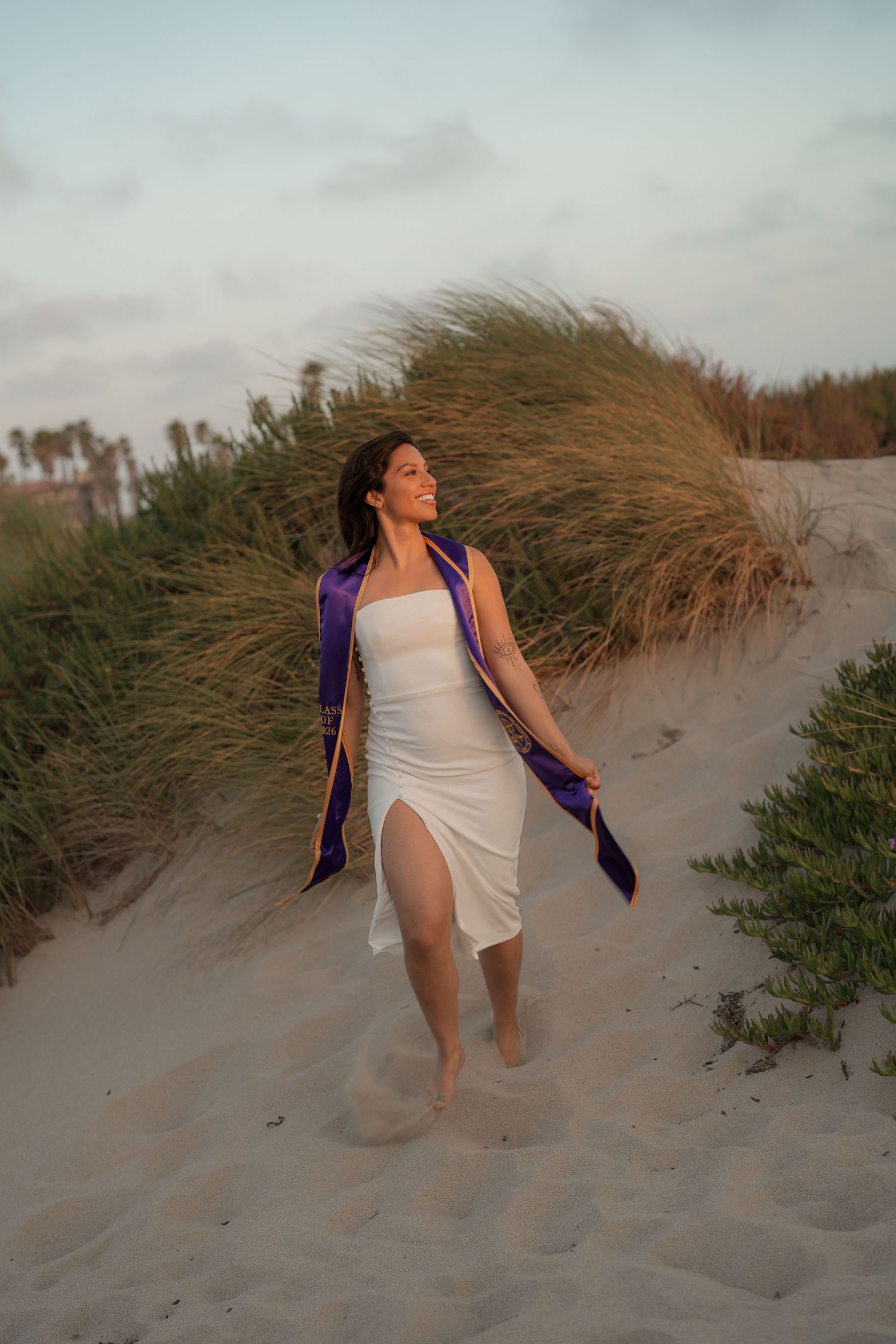 Graduate in a white dress with a purple sash walking barefoot through sand dunes and beach grass at golden hour.