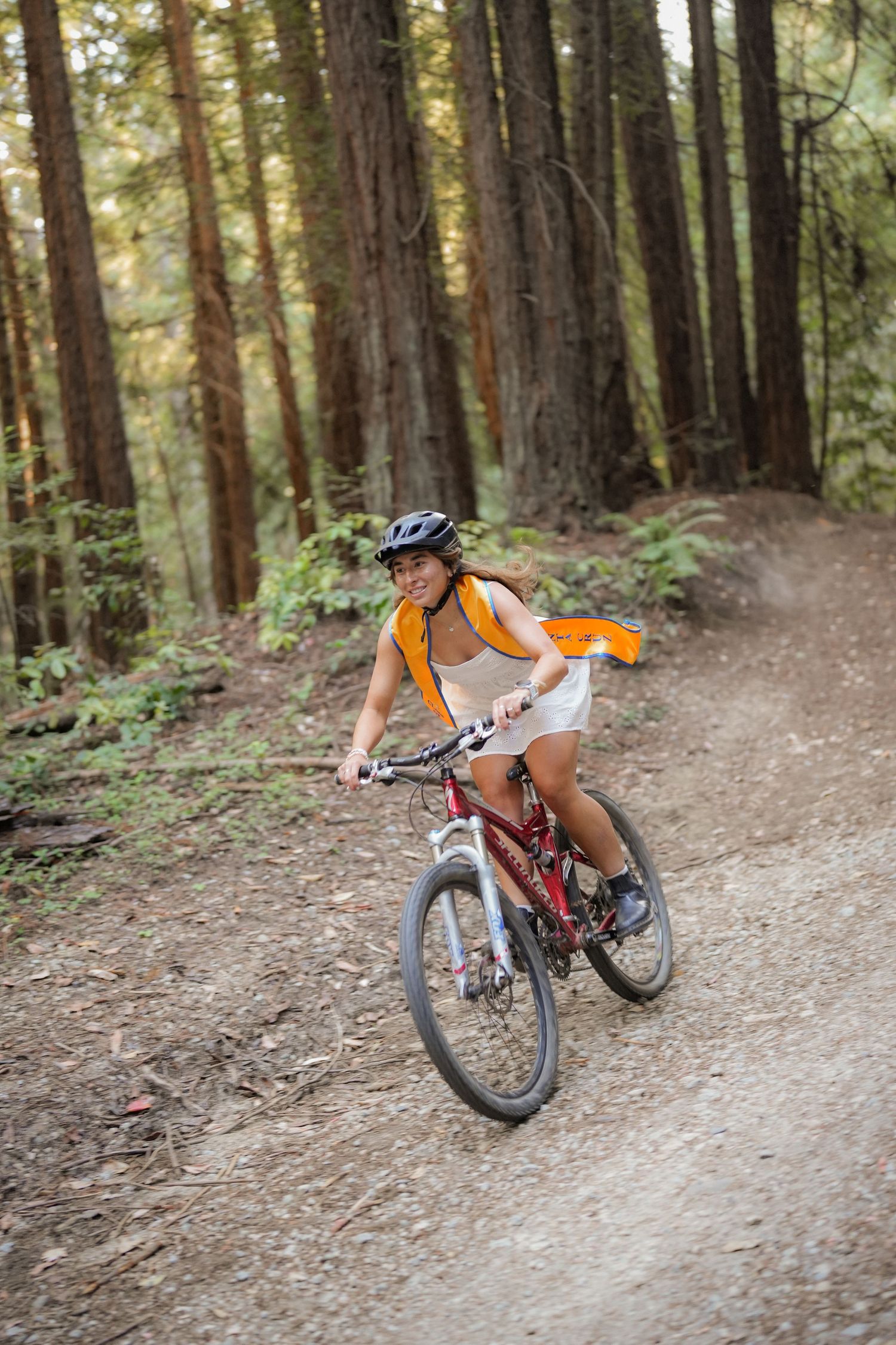 A woman in a helmet and orange vest rides a red mountain bike along a gravel forest trail beneath towering redwoods.