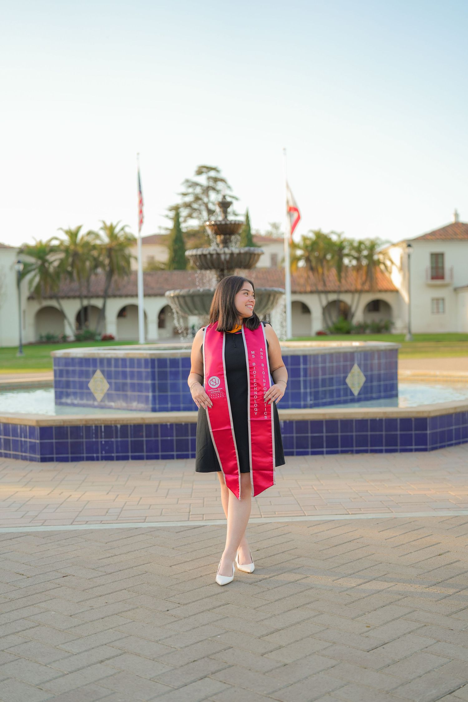 A graduate in a red sash and black dress stands before a blue tiled fountain with flags and Spanish-style buildings behind her.