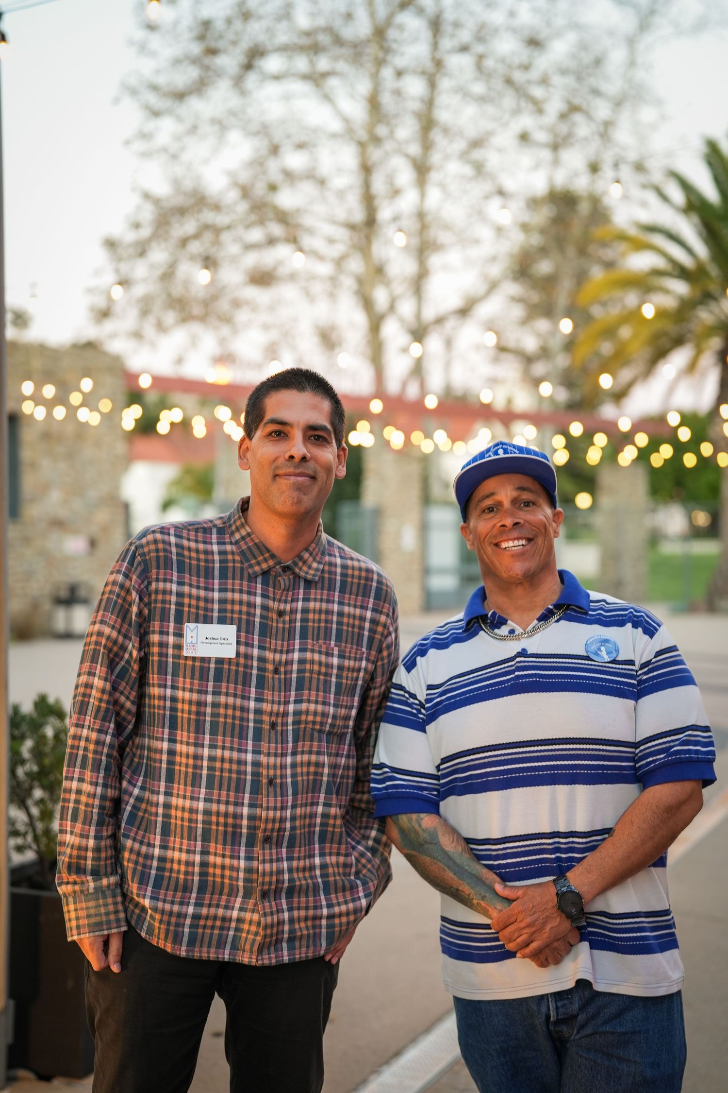 Two smiling men stand side by side outdoors at dusk with string lights and trees glowing behind them.